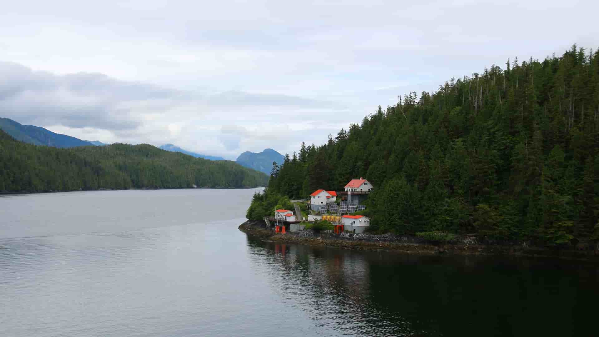 A small cluster of red-roofed buildings on a rocky shoreline in the Queen Charlotte Sound, Canada, surrounded by dense green evergreen forests and mountains under a cloudy sky.