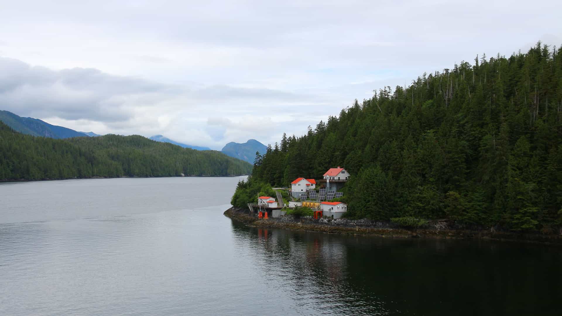 A small cluster of red-roofed buildings on a rocky shoreline in the Queen Charlotte Sound, Canada, surrounded by dense green evergreen forests and mountains under a cloudy sky.