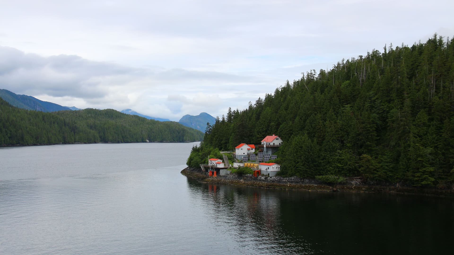 A small cluster of red-roofed buildings on a rocky shoreline in the Queen Charlotte Sound, Canada, surrounded by dense green evergreen forests and mountains under a cloudy sky.