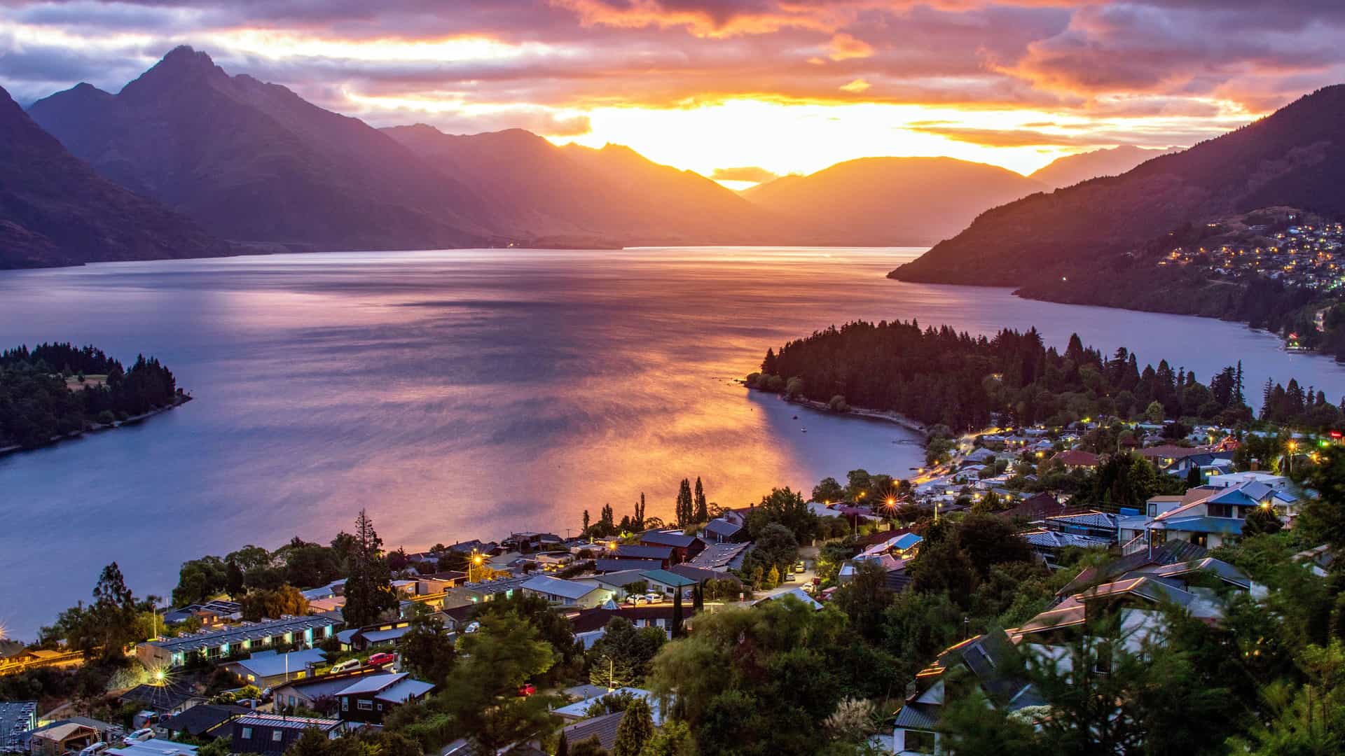 A panoramic view of Queenstown, New Zealand, at sunset, showing the town's lights twinkling along the shores of Lake Wakatipu. The sky is filled with vibrant orange, pink, and purple clouds that are reflected on the tranquil surface of the lake, which is surrounded by majestic, dark mountains.