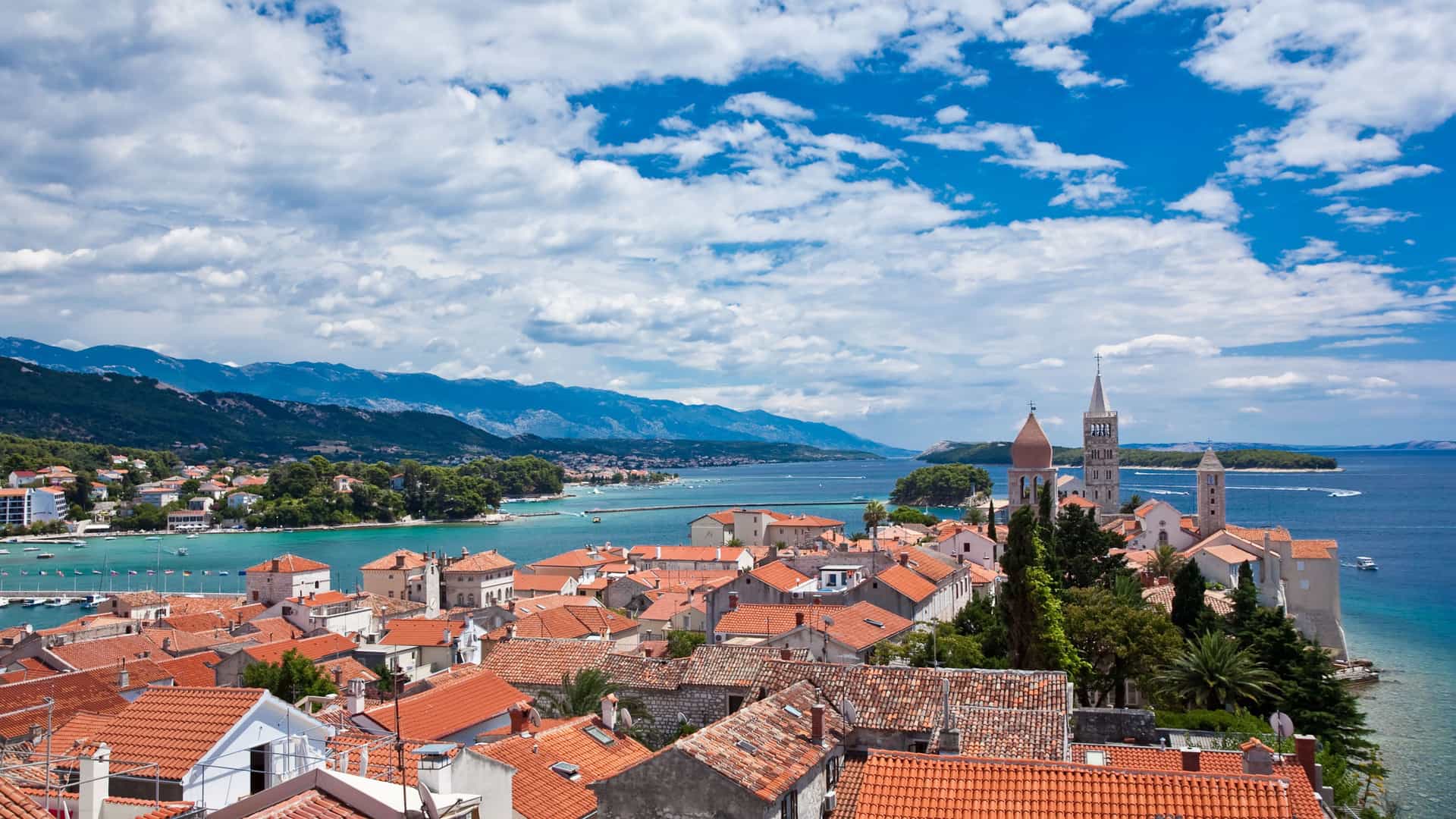 A scenic aerial view of Rab Island, Croatia, showing a charming old town with red-tiled roofs, four bell towers, and a marina next to the turquoise-blue Adriatic Sea.