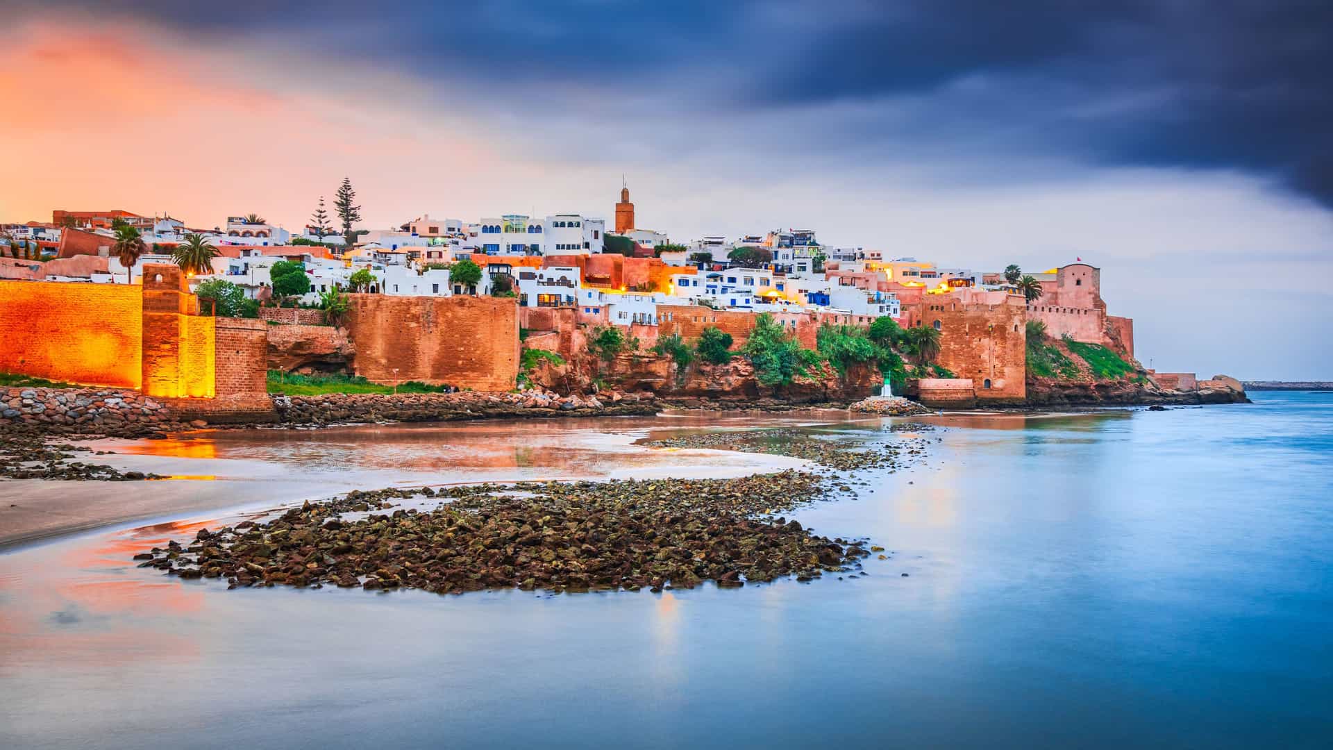 "A scenic view of the Kasbah of the Udayas in Rabat, Morocco, at dusk. The ancient, fortified walls and white-and-blue buildings are warmly lit, overlooking a calm body of water that reflects the vibrant orange, purple, and blue hues of the sunset sky. "