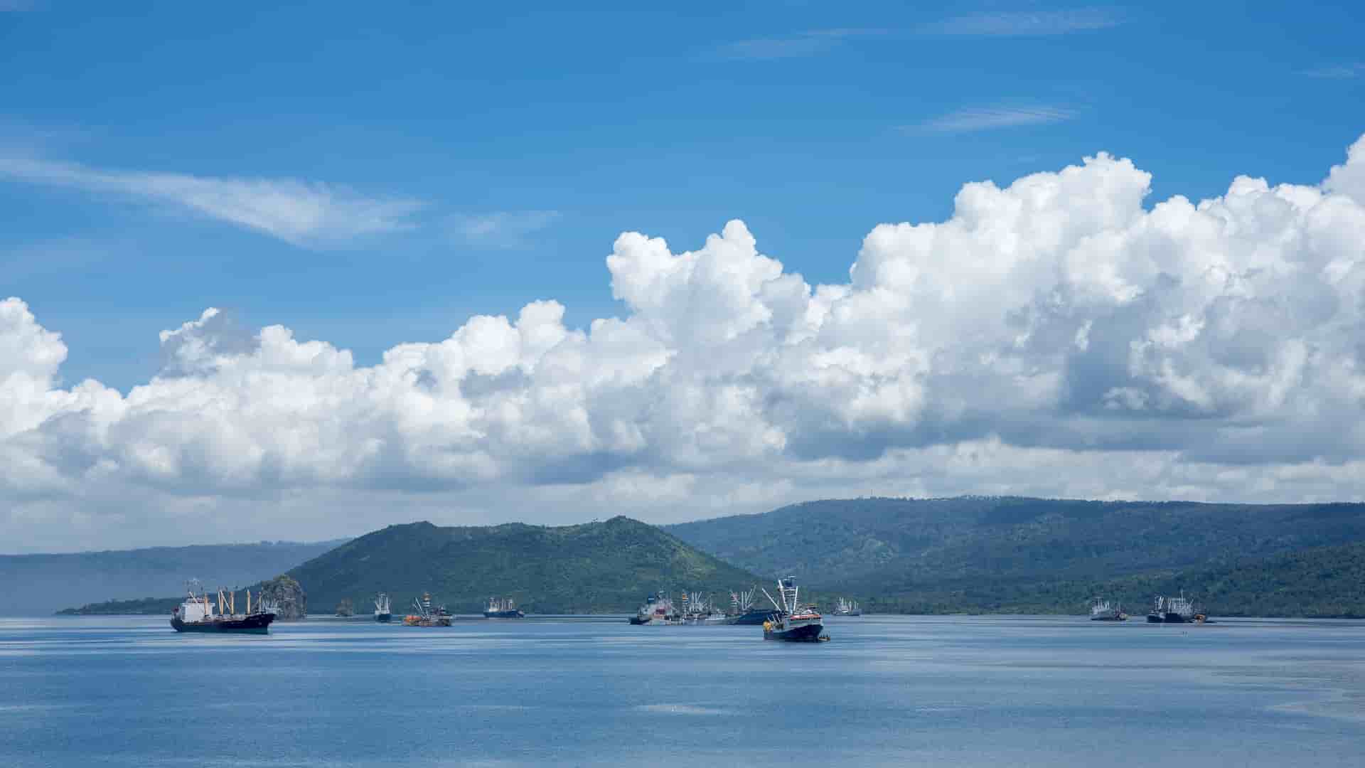 A serene view of Simpson Harbour in Rabaul, Papua New Guinea, with multiple cargo and fishing boats anchored in the calm water with an active volcano in the distance under a blue sky with fluffy white clouds.