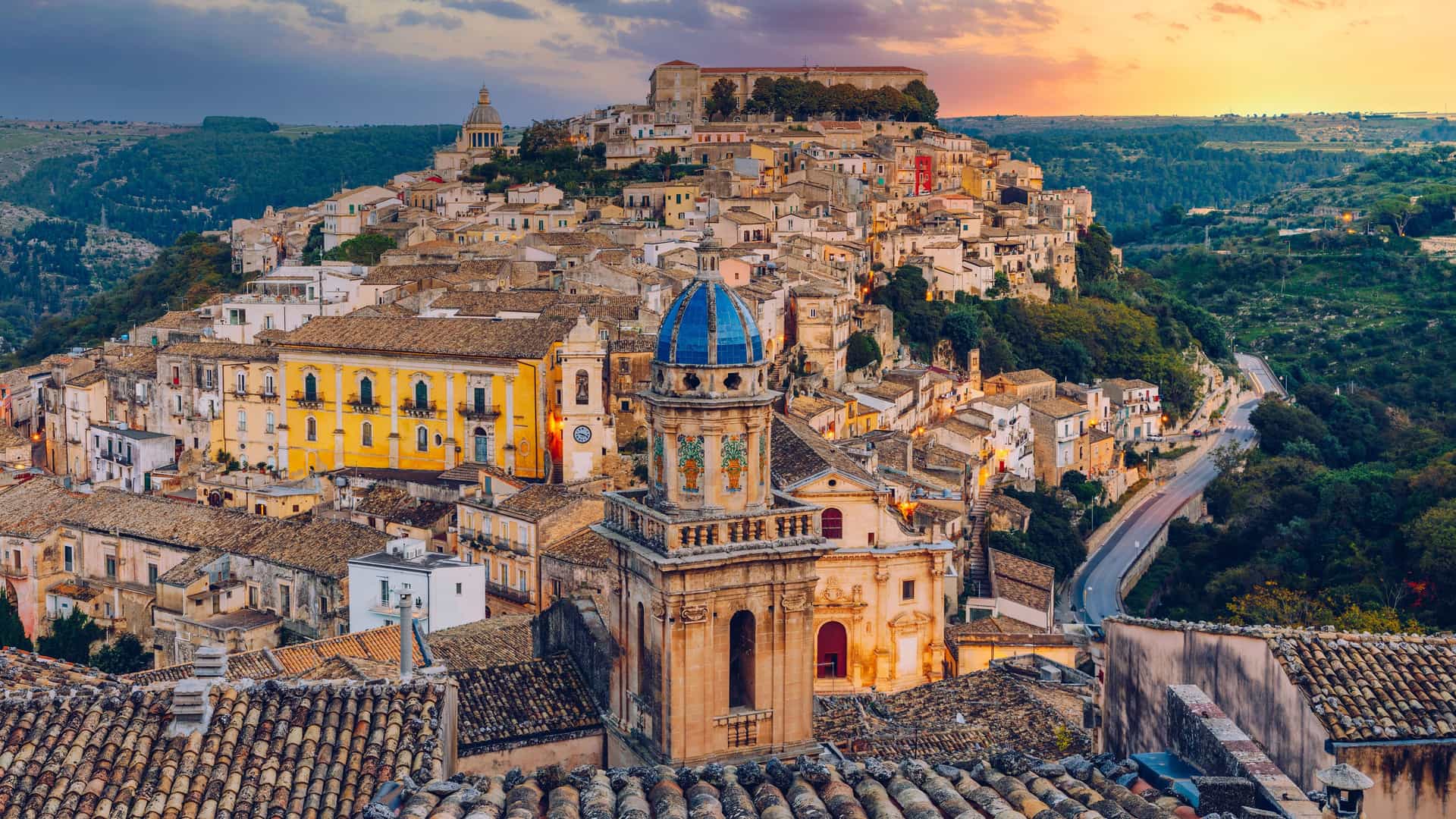 "An elevated view of the historic Baroque town of Ragusa Ibla in Sicily, Italy, showing a dense cluster of buildings and terracotta rooftops on a steep hillside. In the foreground, a prominent church with a striking blue dome and clock tower stands out, with the warm colors of a sunset filling the sky above. "
