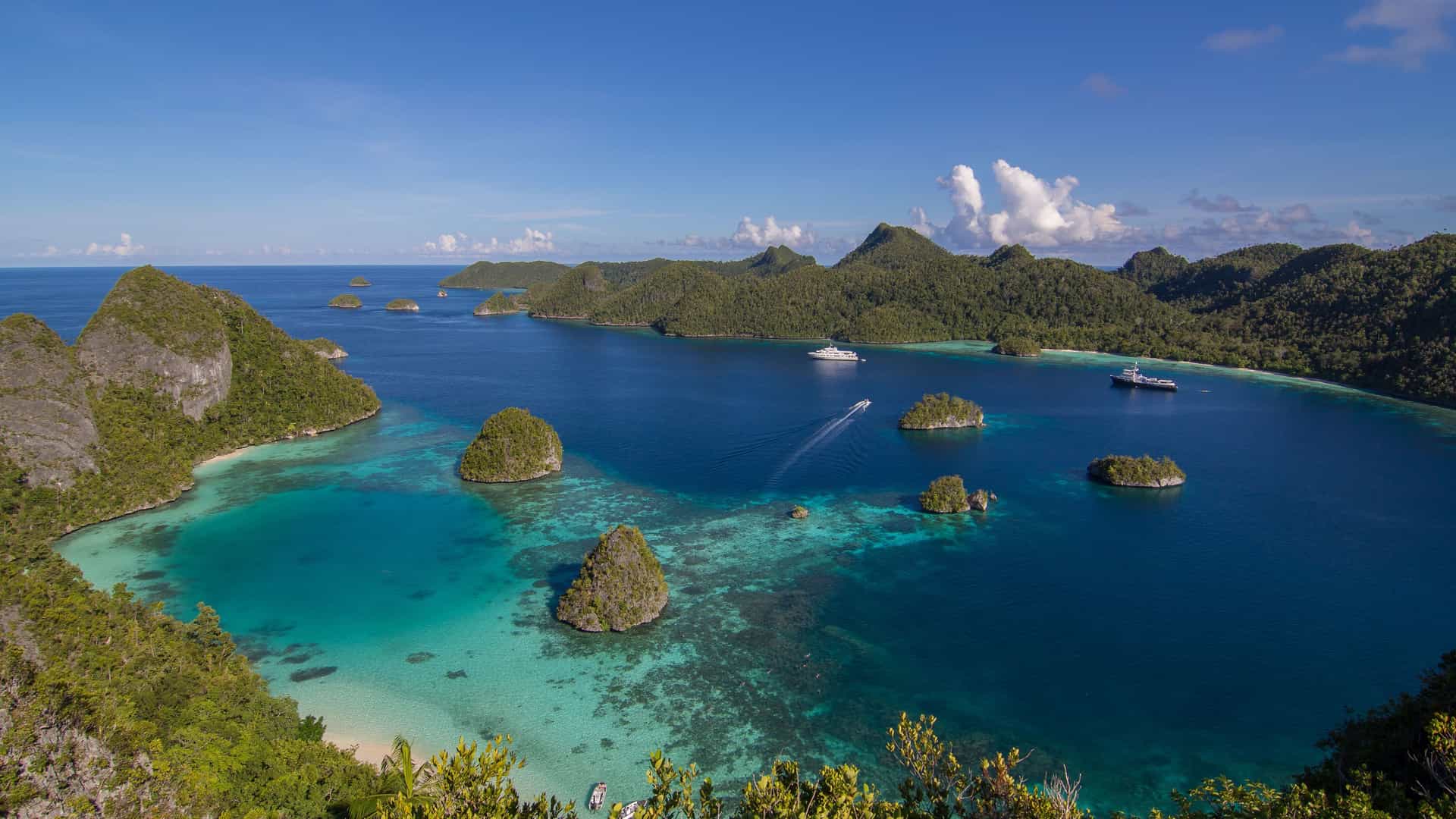A wide-angle shot of the stunning Raja Ampat islands, Indonesia, showing a series of small, green-covered limestone karsts rising from a crystal-clear turquoise lagoon.