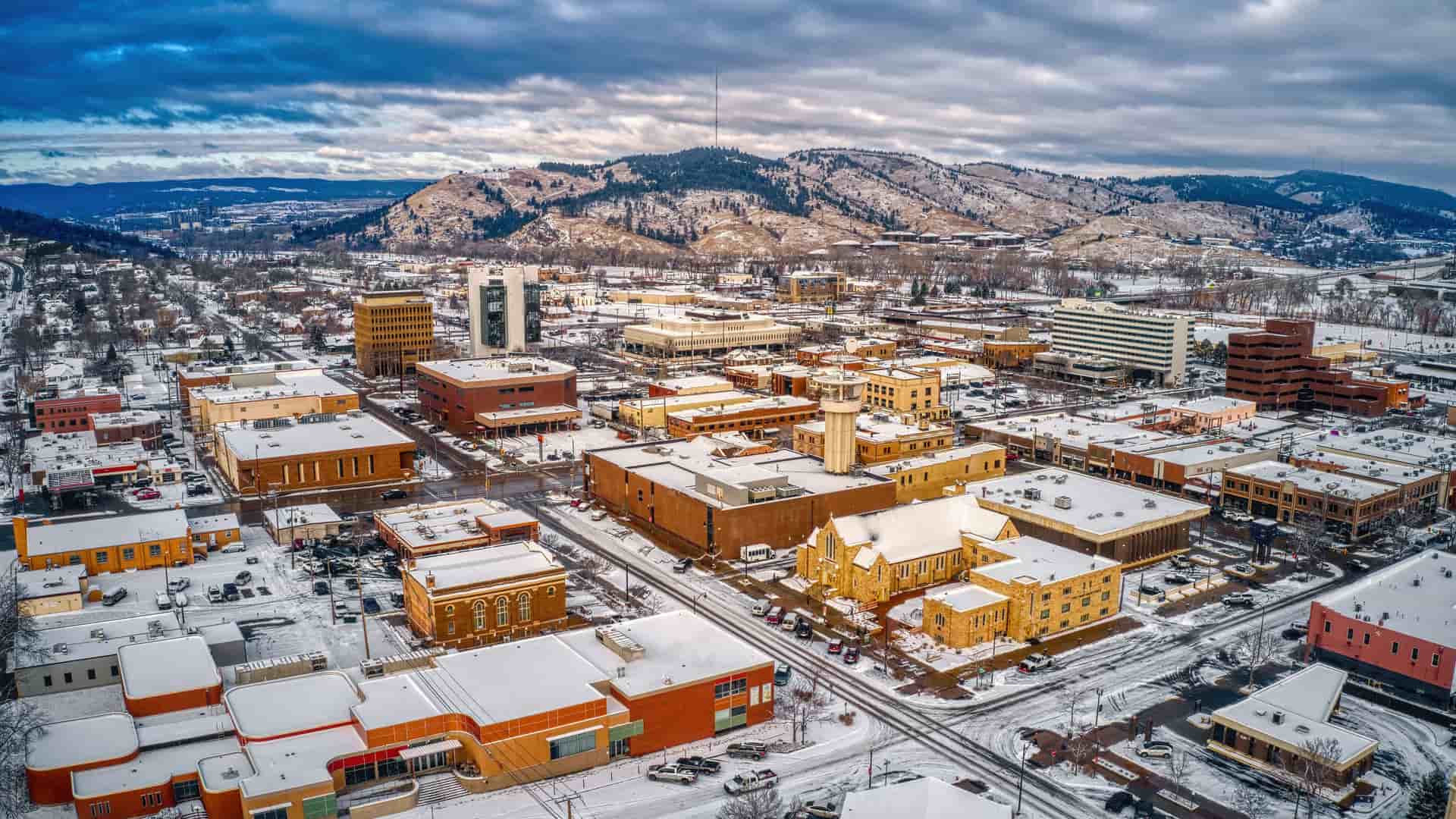 "An aerial view of downtown Rapid City, South Dakota, covered in a light blanket of snow during winter. The buildings, a mix of modern and older low-rises, have rooftops dusted with white, and the streets are visible as dark lines. The city is nestled against the backdrop of snow-covered Black Hills, with evergreen trees and patches of white snow on the slopes under a dramatic, cloudy sky. "