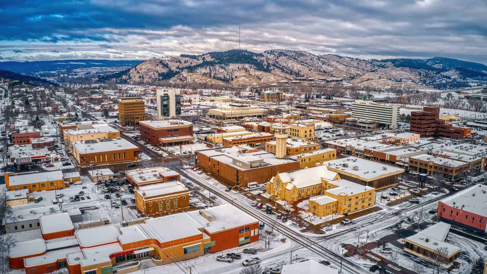 "An aerial view of downtown Rapid City, South Dakota, covered in a light blanket of snow during winter. The buildings, a mix of modern and older low-rises, have rooftops dusted with white, and the streets are visible as dark lines. The city is nestled against the backdrop of snow-covered Black Hills, with evergreen trees and patches of white snow on the slopes under a dramatic, cloudy sky. "