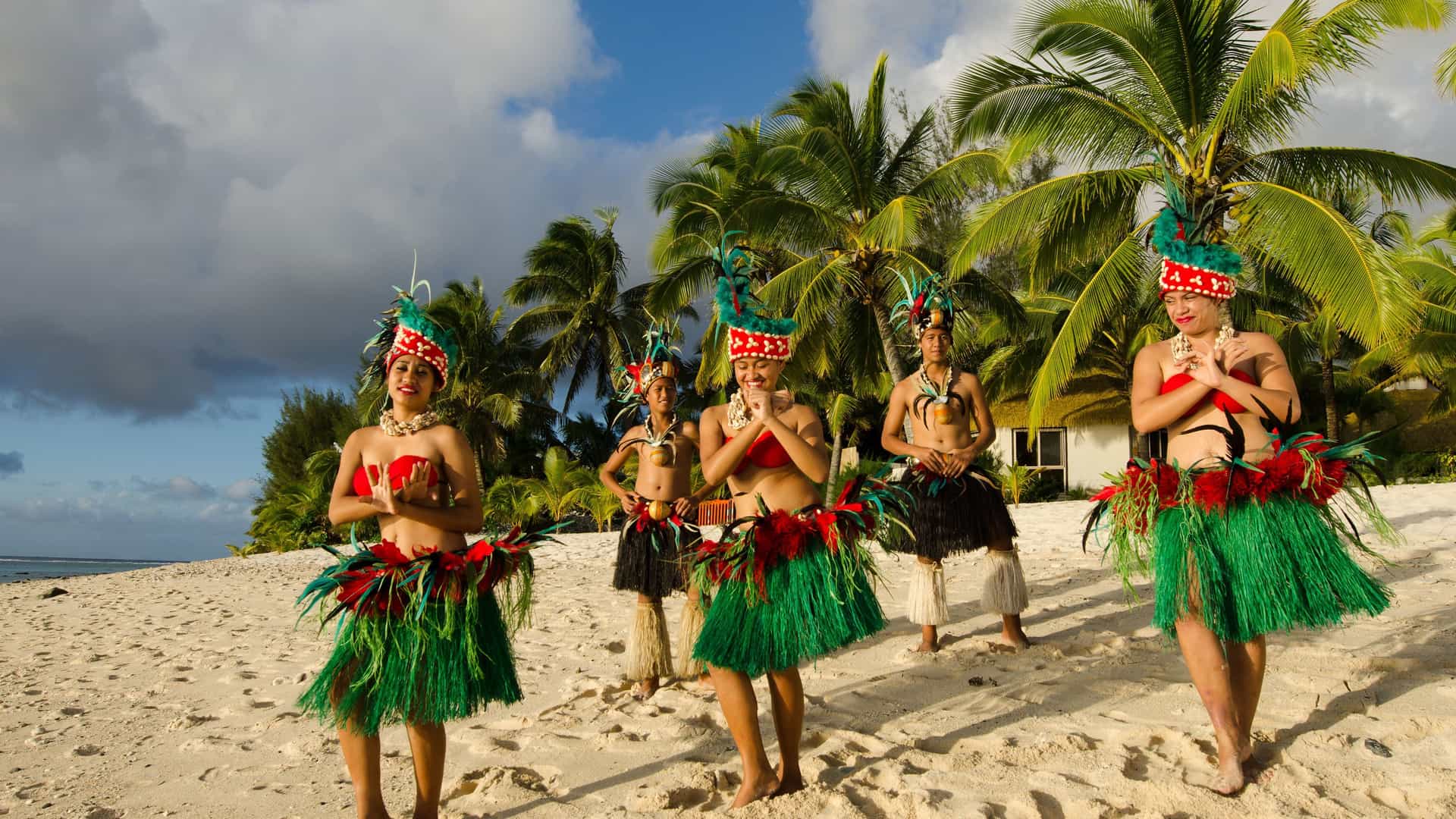 A group of five Cook Islands women in traditional dress with green skirts, red feathers, and colorful headdresses perform a dance on a sandy Rarotonga beach surrounded by palm trees.