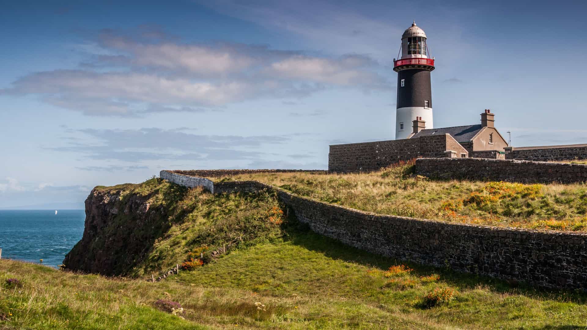 "A scenic view of the Rathlin West Lighthouse on Rathlin Island, Northern Ireland. The black and white lighthouse, with its red top, is situated on a lush green cliff, with low, winding stone walls defining the grounds. The lighthouse overlooks the deep blue sea under a partly cloudy sky. "