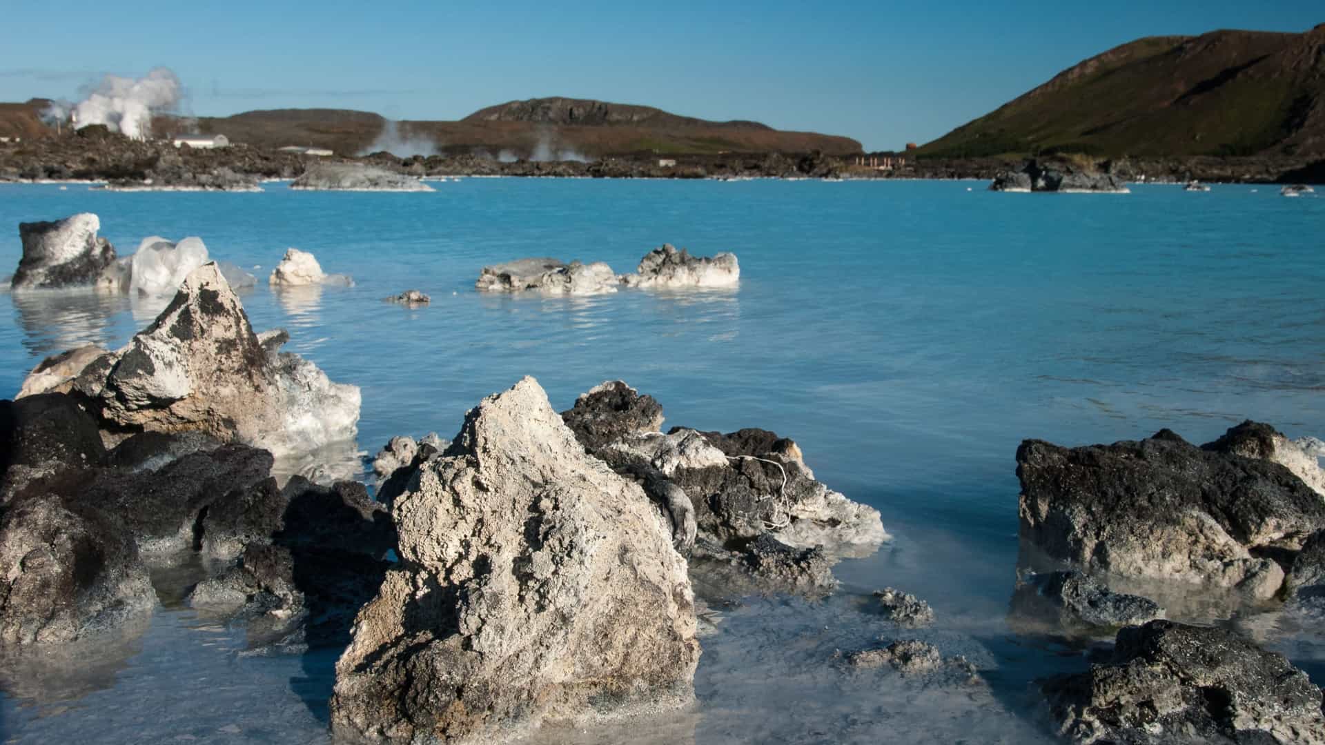Geothermal activity sends steam plumes into the air behind black lava rocks and light blue, mineral-rich water in Raudanes, Iceland.