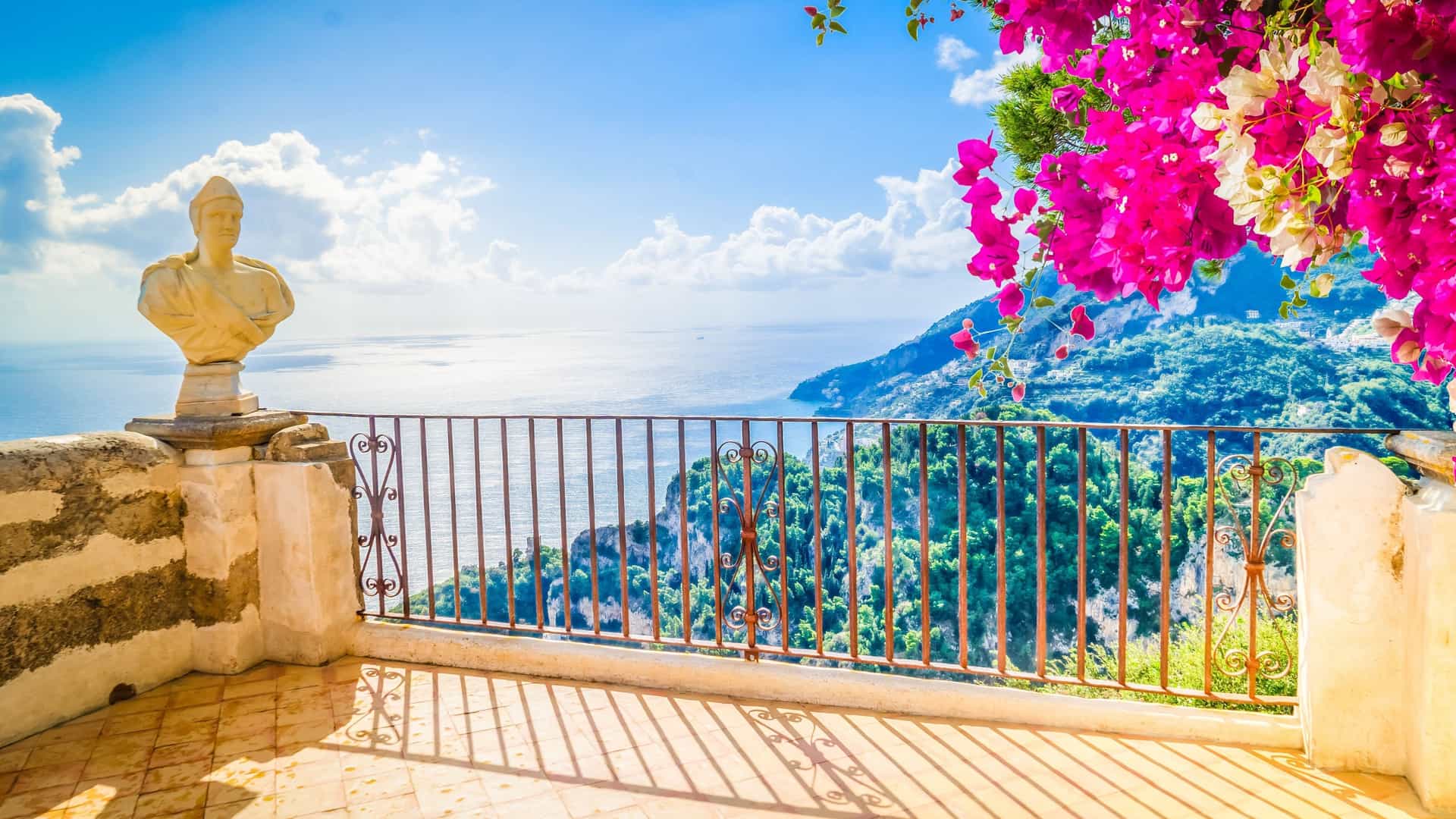 A sunny, tiled terrace overlooking the Amalfi Coast features a classical bust and bright pink bougainvillea flowers.