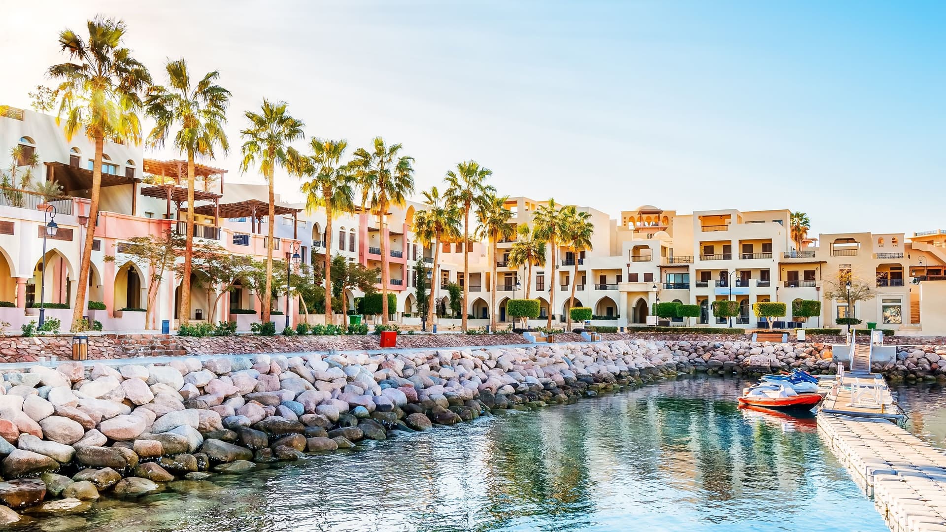 A beautiful view of the marina in Aqaba, Jordan, featuring traditional-style buildings and palm trees lining a stone-paved waterfront along the Red Sea.