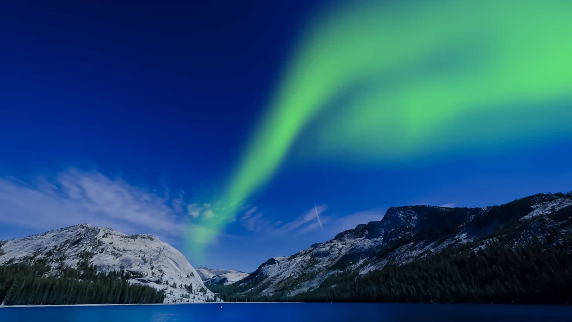 A vibrant display of the green Northern Lights, or Aurora Borealis, illuminating the dark blue sky over a snow-capped mountain and calm water on Resolution Island.