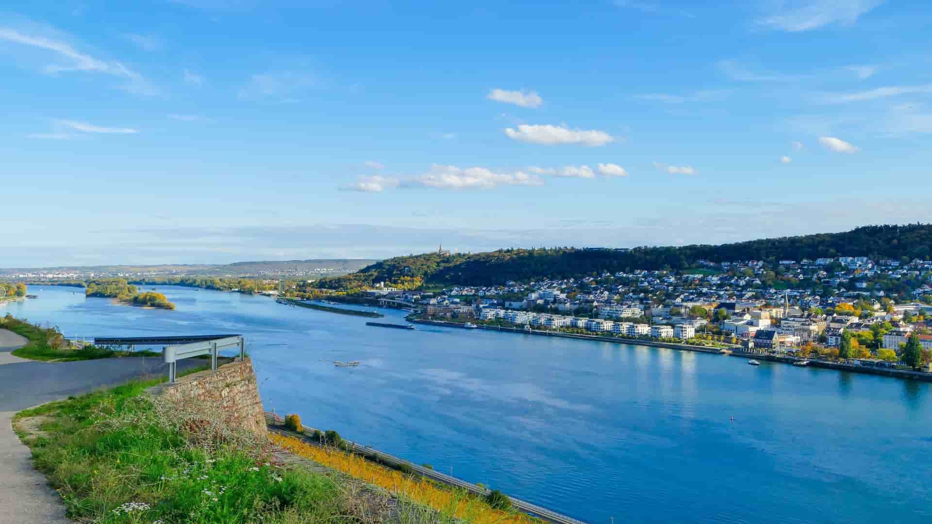 An elevated view of the wide, blue Rhine River flowing through a large valley, with a town nestled on the far bank.