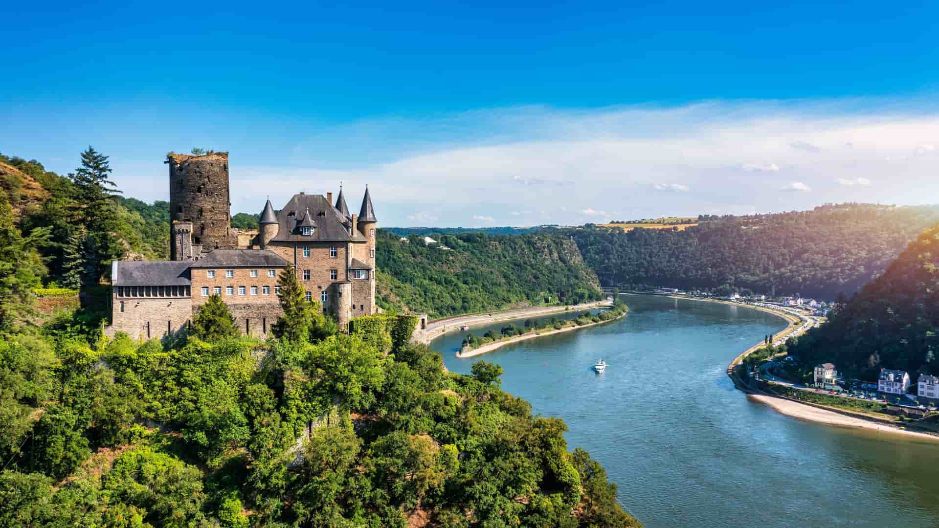 "A scenic view of a medieval German castle with turrets and towers, perched on a hill overlooking the Rhine River, a popular cruise destination in Rhineland. "