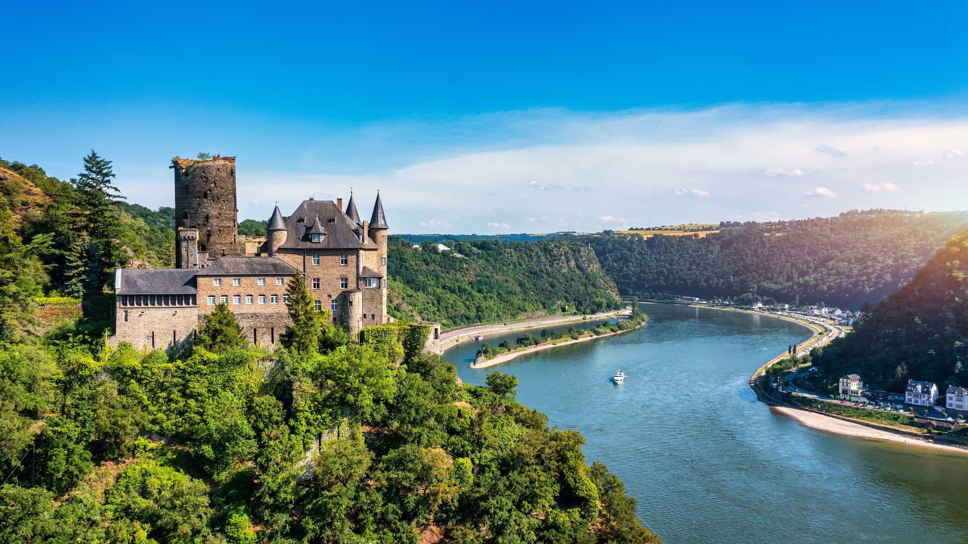 "A scenic view of a medieval German castle with turrets and towers, perched on a hill overlooking the Rhine River, a popular cruise destination in Rhineland. "