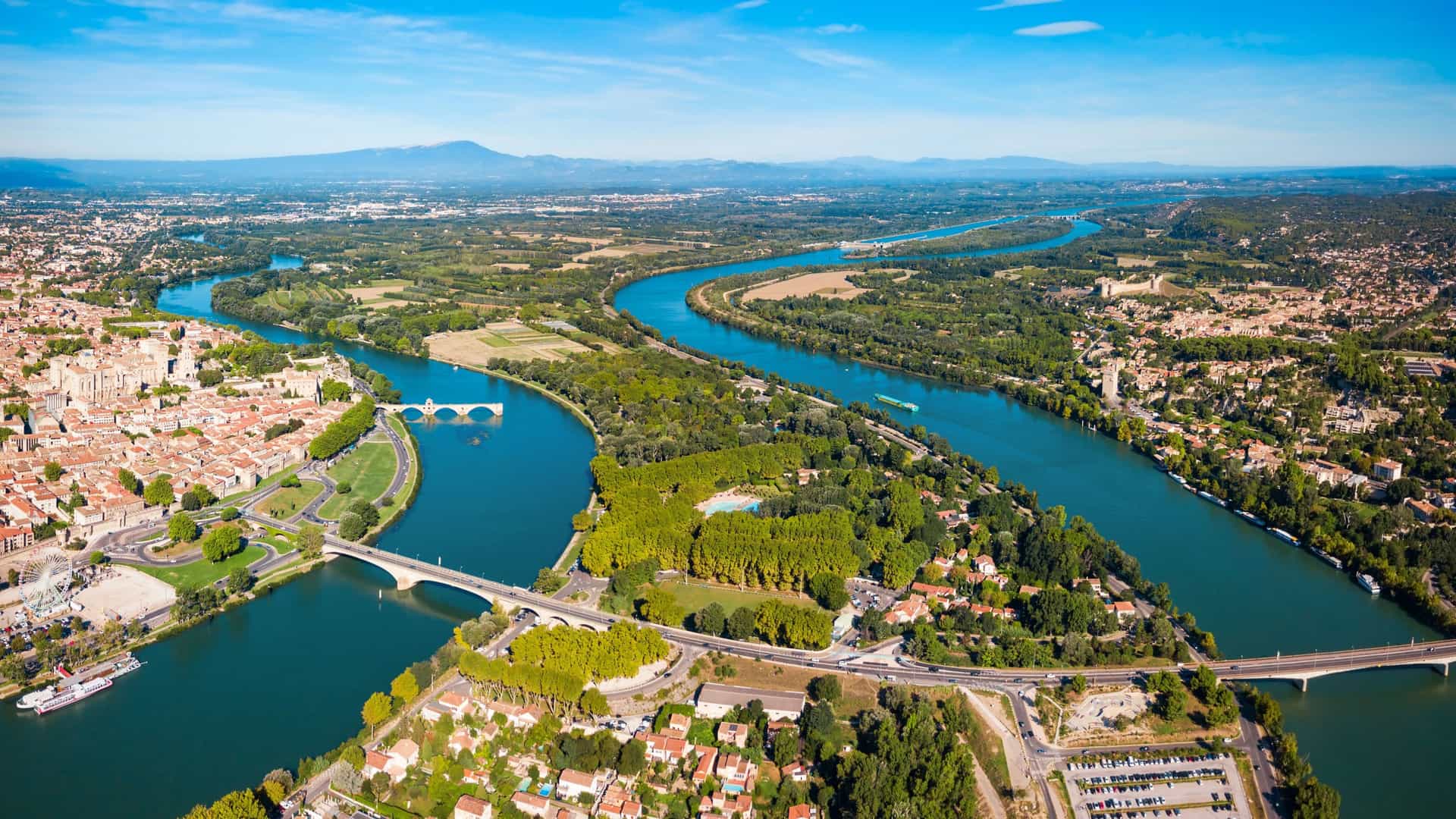 An aerial view of Avignon, France, showing the famous Pont d'Avignon bridge and the city's historic buildings along the wide, curving Rhône River on a clear, sunny day.