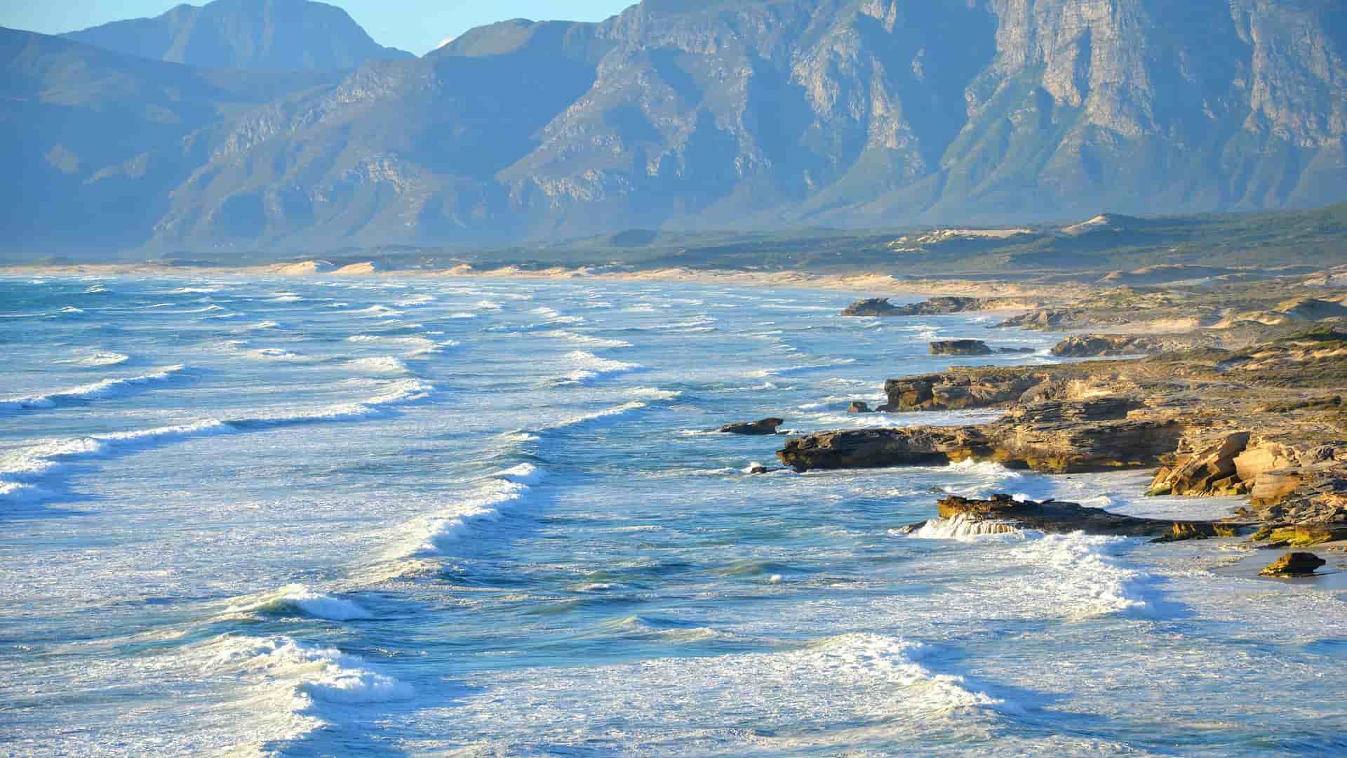 A dramatic view of the rugged coastline in Richards Bay, South Africa, with waves crashing against a rocky shore and majestic mountains in the distant background.