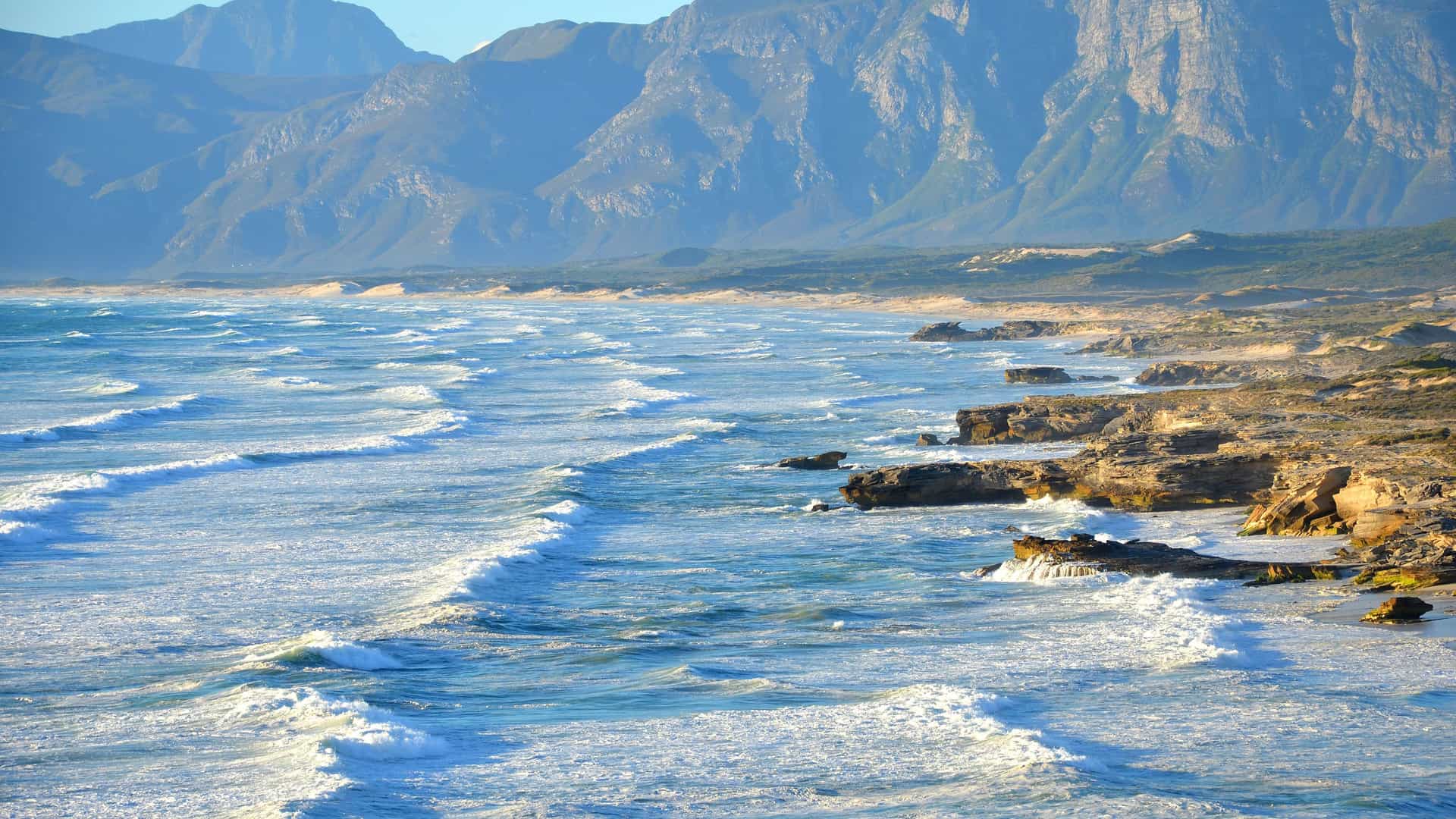 A dramatic view of the rugged coastline in Richards Bay, South Africa, with waves crashing against a rocky shore and majestic mountains in the distant background.
