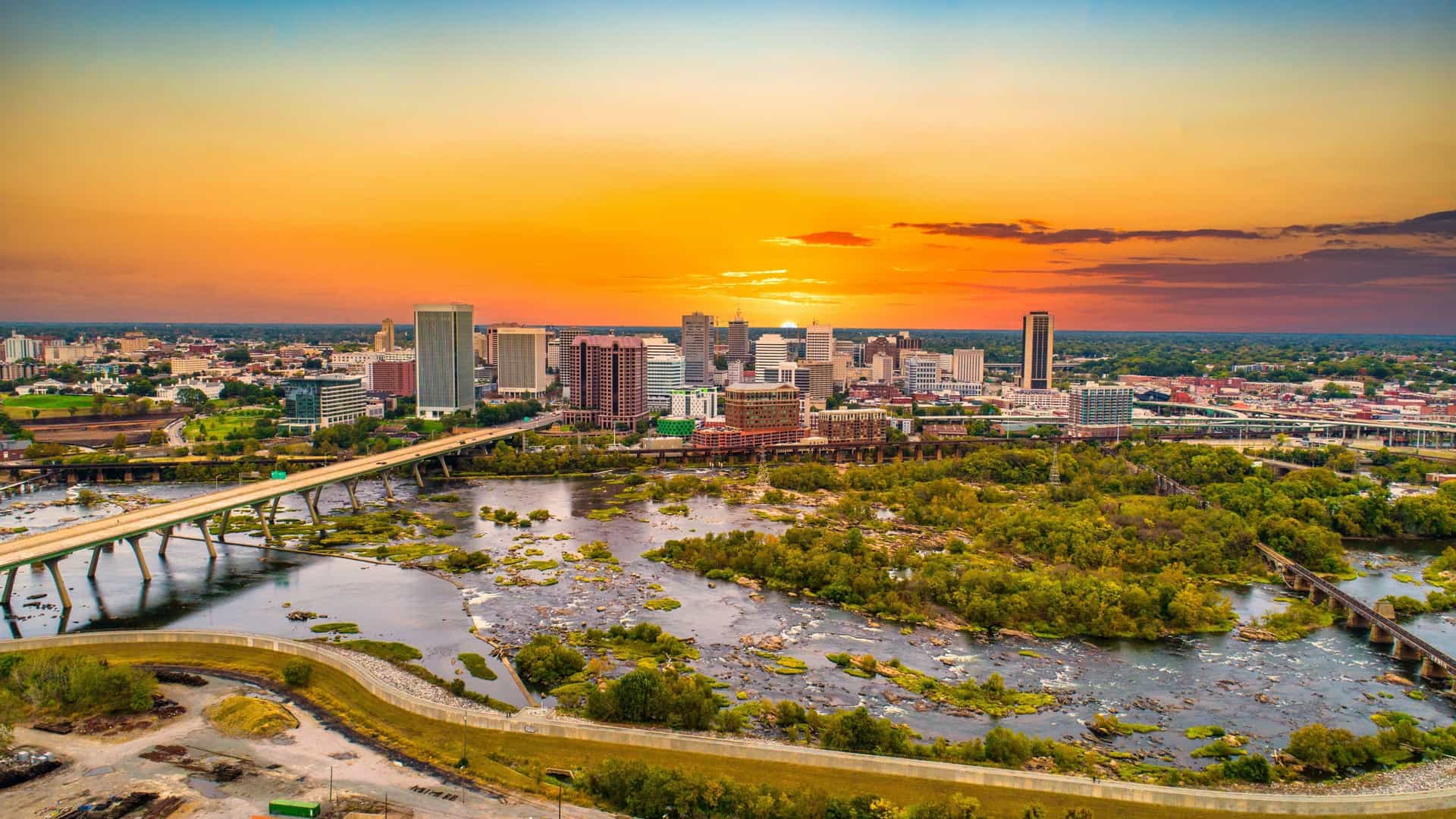 A high-angle drone view of the downtown Richmond, Virginia, USA skyline, showcasing a cluster of modern high-rise buildings and older brick structures. The cityscape is situated along the banks of the James River, with a bridge or railroad trestle visible crossing the water in the foreground or midground. The image is taken during the day, likely featuring a clear or partly cloudy sky.