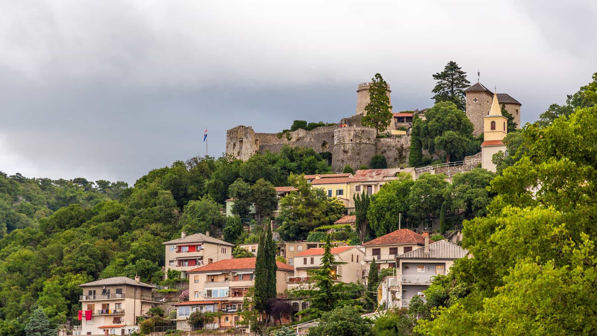 A beautiful view of Trsat Castle, a medieval fortress perched atop a hill overlooking the city of Rijeka, Croatia, with residential buildings and lush green trees below.
