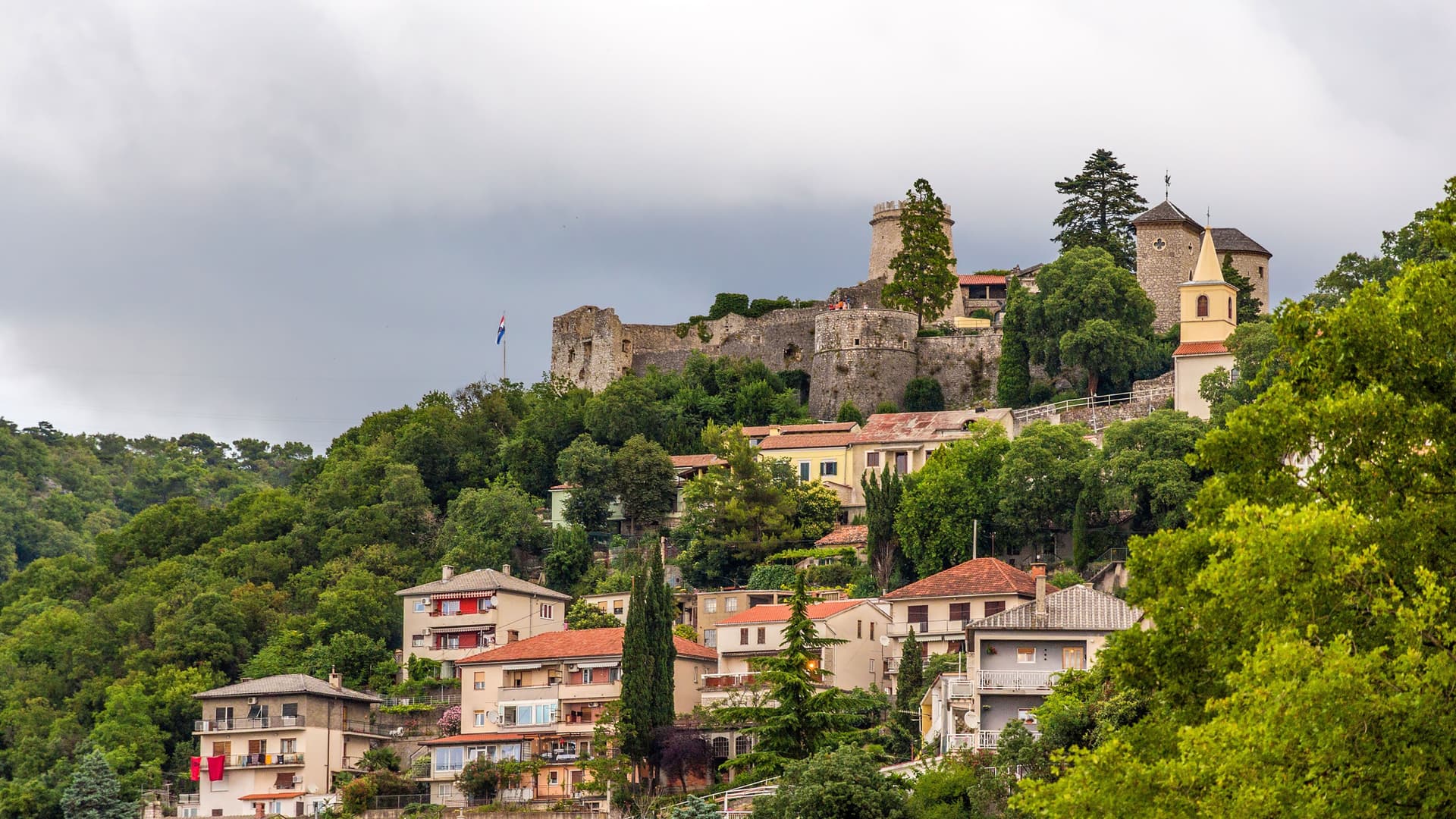A beautiful view of Trsat Castle, a medieval fortress perched atop a hill overlooking the city of Rijeka, Croatia, with residential buildings and lush green trees below.