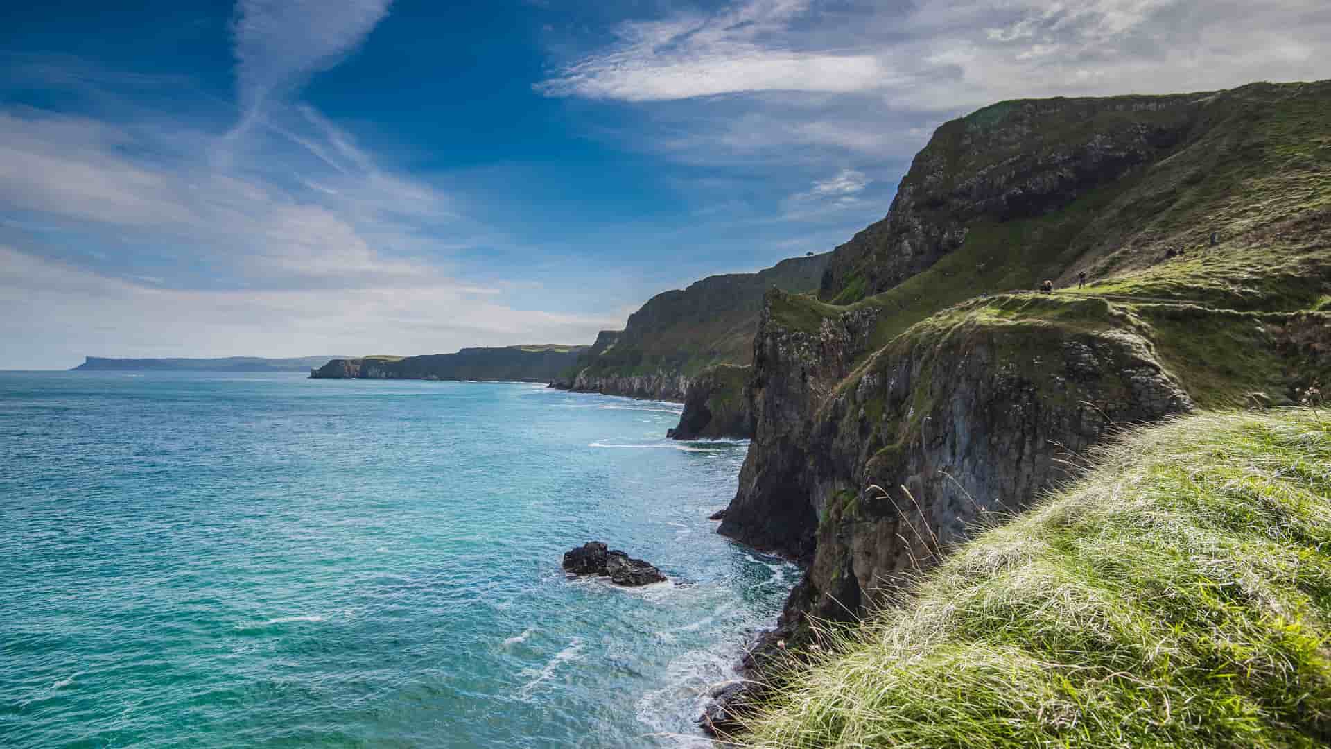 A dramatic view of the rugged coastline in Ringaskiddy, Ireland, featuring green, grassy cliffs, the deep blue Atlantic Ocean, and a vibrant sky with wispy clouds.