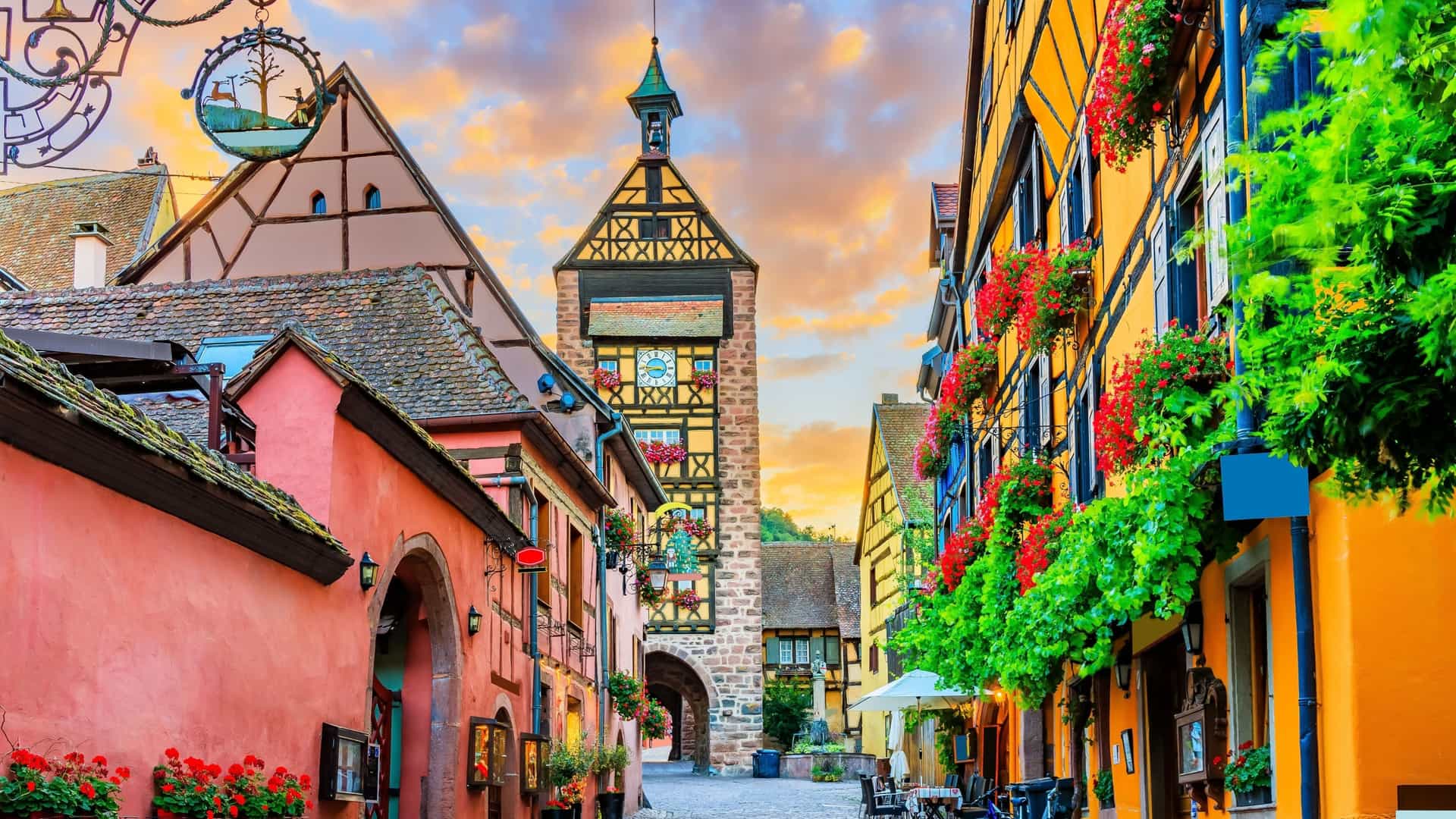 A charming view of the Dolder Tower and cobblestone street in Riquewihr, France, featuring colorful, half-timbered buildings adorned with flower baskets and a dramatic sunset sky.