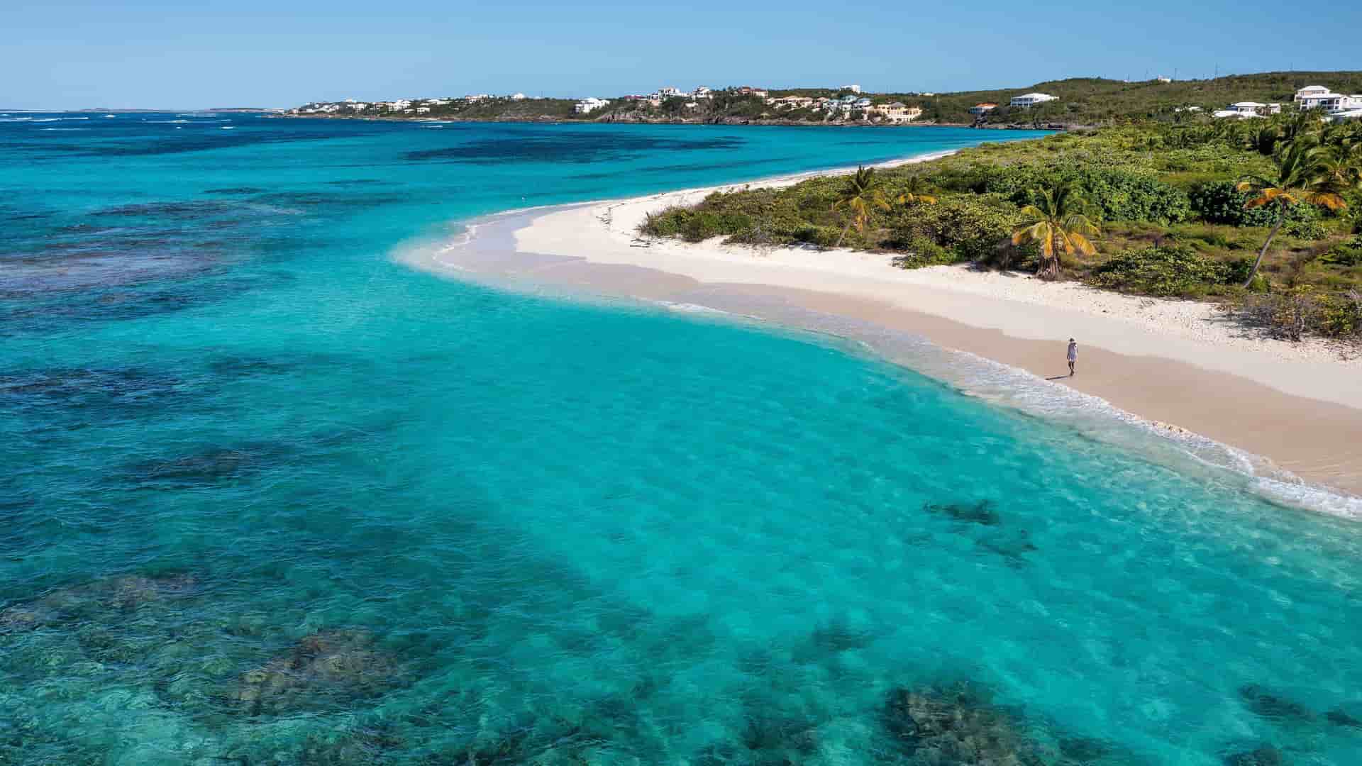 An aerial view of Road Bay, Anguilla, showing a pristine white sand beach, lush green vegetation, and the stunningly clear turquoise water of the Caribbean Sea.