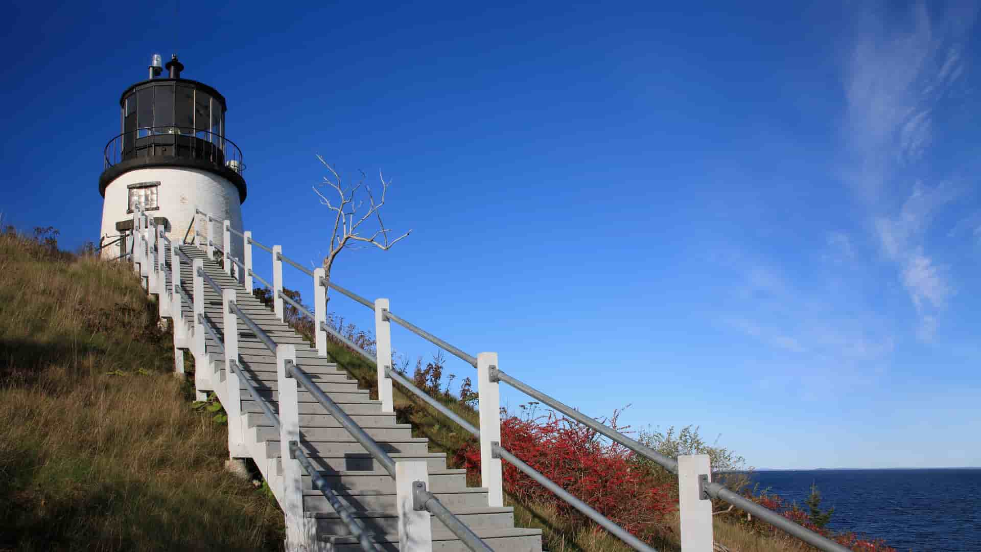 A view of the Owl's Head Lighthouse in Rockland, Maine, with a long staircase leading up to the historic white lighthouse, set against a bright blue sky and the Atlantic Ocean.