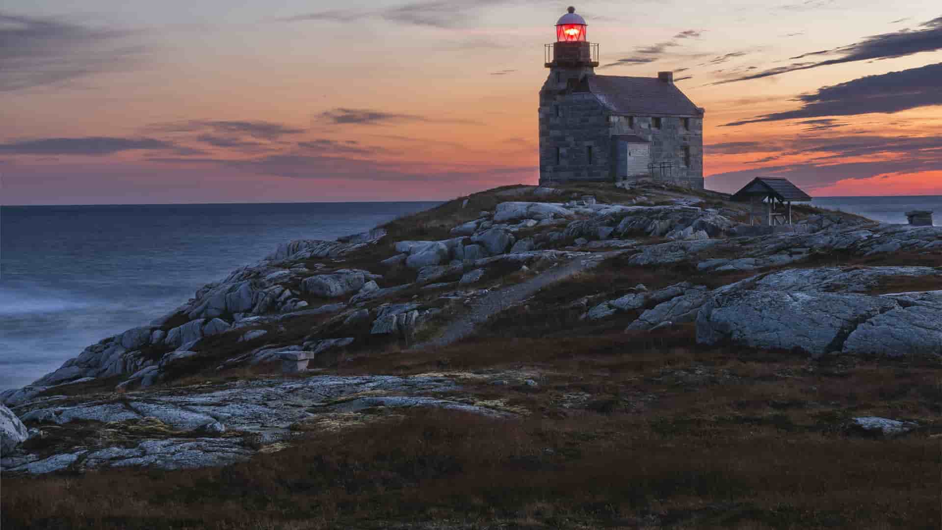 A stunning sunset view of a historic stone lighthouse on Rose Island in Newfoundland and Labrador, with its light glowing as the sun sets over the Atlantic Ocean.