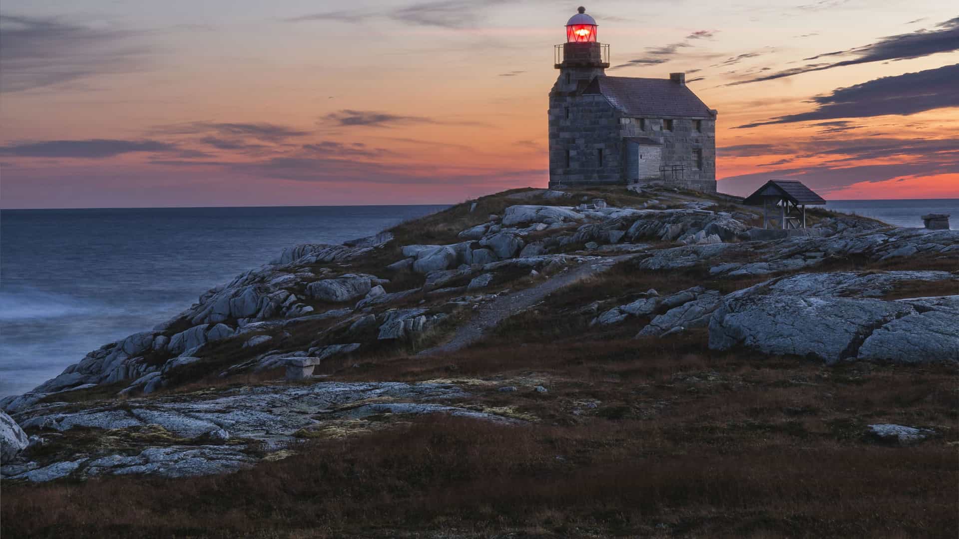 A stunning sunset view of a historic stone lighthouse on Rose Island in Newfoundland and Labrador, with its light glowing as the sun sets over the Atlantic Ocean.