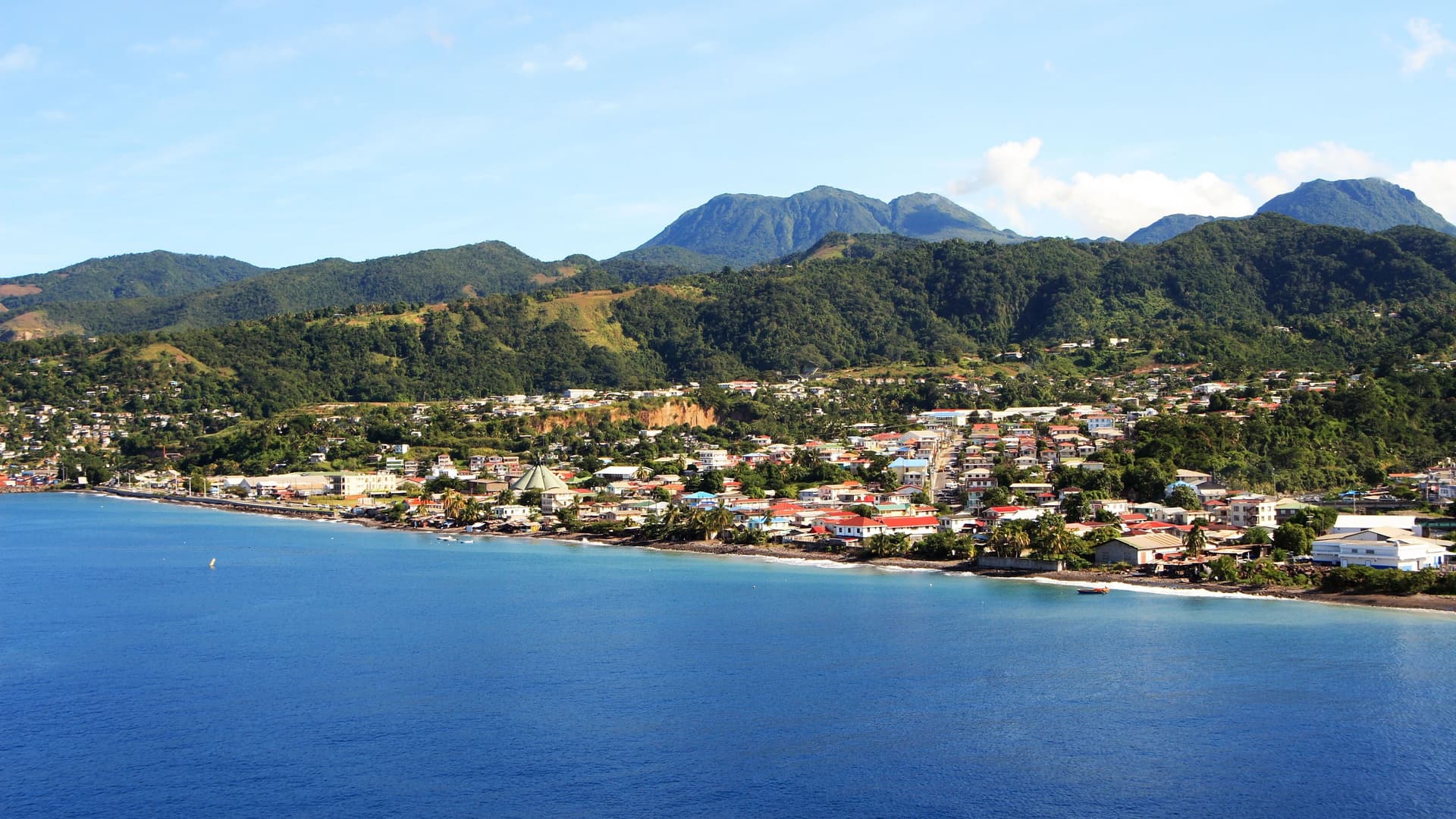A beautiful panoramic view of Roseau, Dominica, with a colorful town nestled between the deep blue Caribbean Sea and a backdrop of lush green, volcanic mountains.