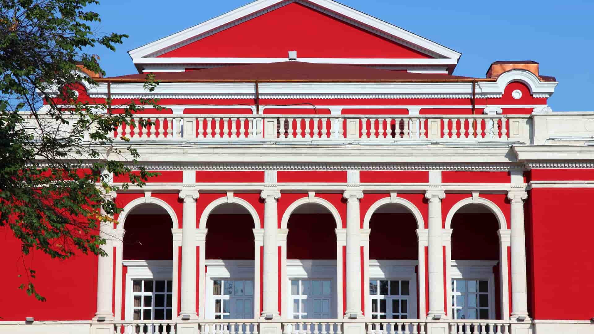 A striking close-up of the facade of the red-and-white neoclassical theater building in Rousse, Bulgaria, with large arched windows and a detailed balcony.