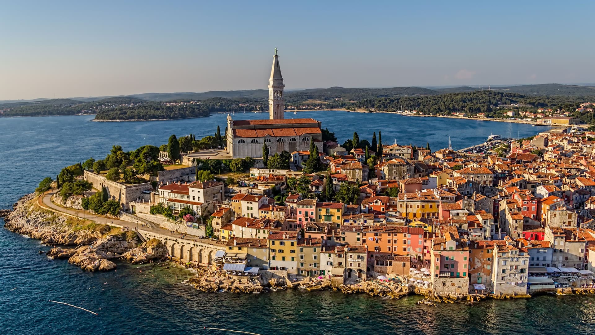 An aerial view of the historic town of Rovinj, Croatia, with its colorful buildings and the bell tower of St. Euphemia's Church dominating the skyline, all perched on a peninsula jutting into the Adriatic Sea.