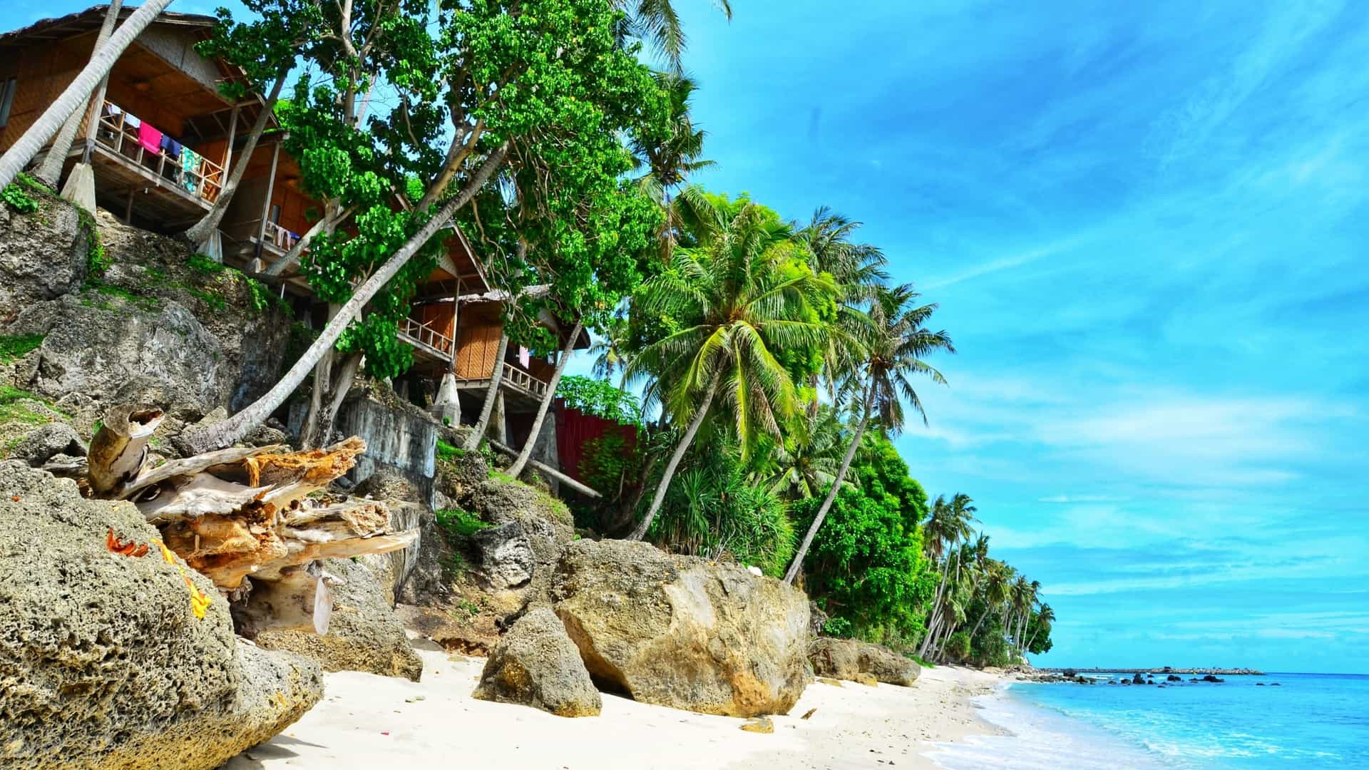 A serene beach scene on Pulau Weh, near Sabang, with a sandy shore and turquoise water, backed by lush green palm trees and wooden huts perched on a rocky cliffside.