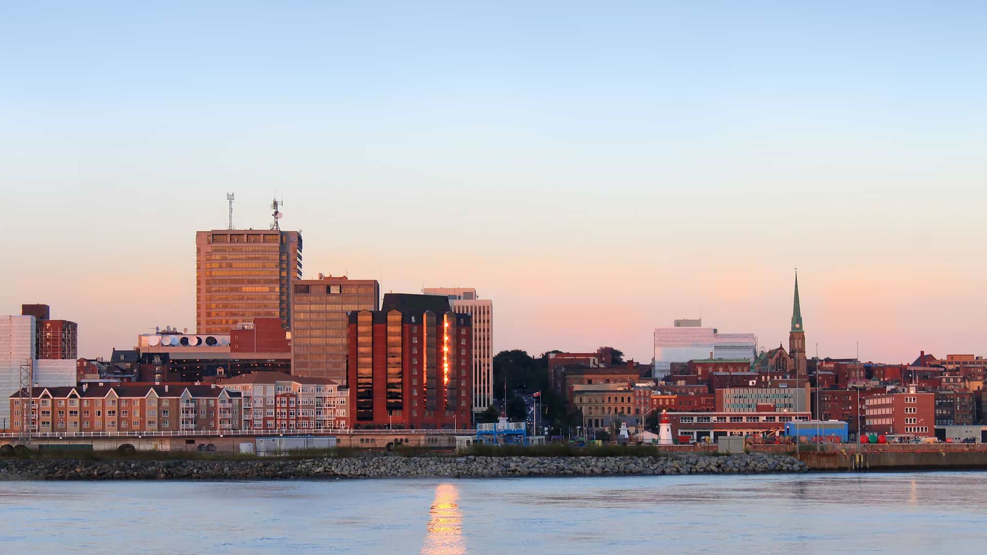 A cityscape view of a small town on the Saguenay River with a mix of modern high-rises and historic buildings with a church steeple, all bathed in the warm light of dusk.