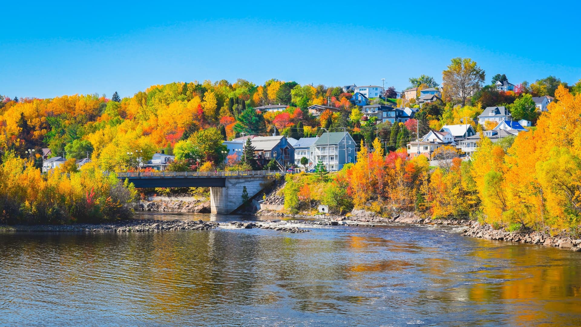 A beautiful autumn landscape in Saguenay with a river flowing under a bridge, surrounded by hillsides adorned with houses and vibrant yellow, orange, and red fall foliage.