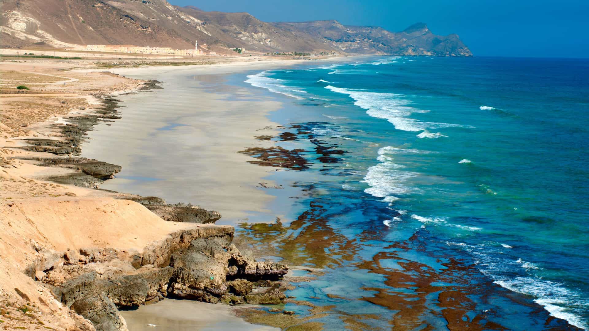 A high-angle view of a long, sandy beach in Salalah, Oman, where gentle ocean waves wash ashore, with rugged, arid mountains forming the coastline under a clear blue sky.