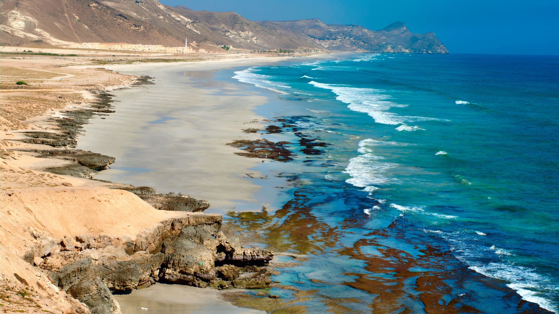 A high-angle view of a long, sandy beach in Salalah, Oman, where gentle ocean waves wash ashore, with rugged, arid mountains forming the coastline under a clear blue sky.