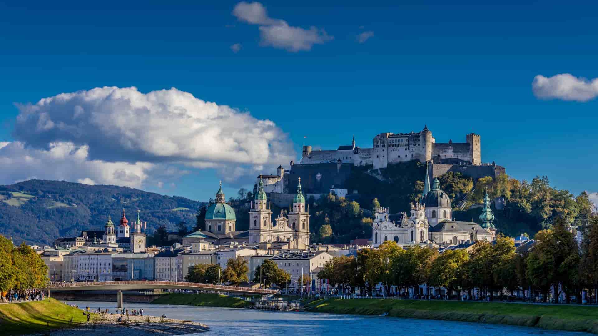 A picturesque view of the historic city of Salzburg, Austria, with the Salzach River in the foreground, the Hohensalzburg Fortress perched on a hill, and a beautiful mix of baroque churches and buildings.