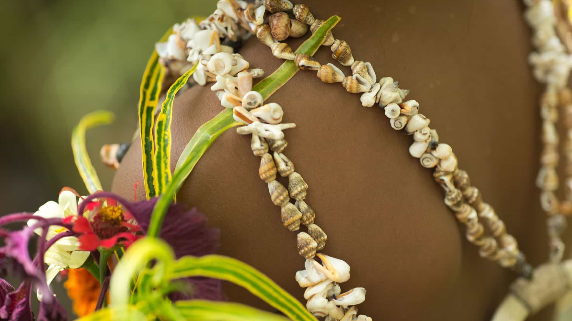 A close-up of a person's shoulder adorned with traditional necklaces made of small seashells and vibrant, colorful flowers and leaves on Samarai Island, Papua New Guinea.