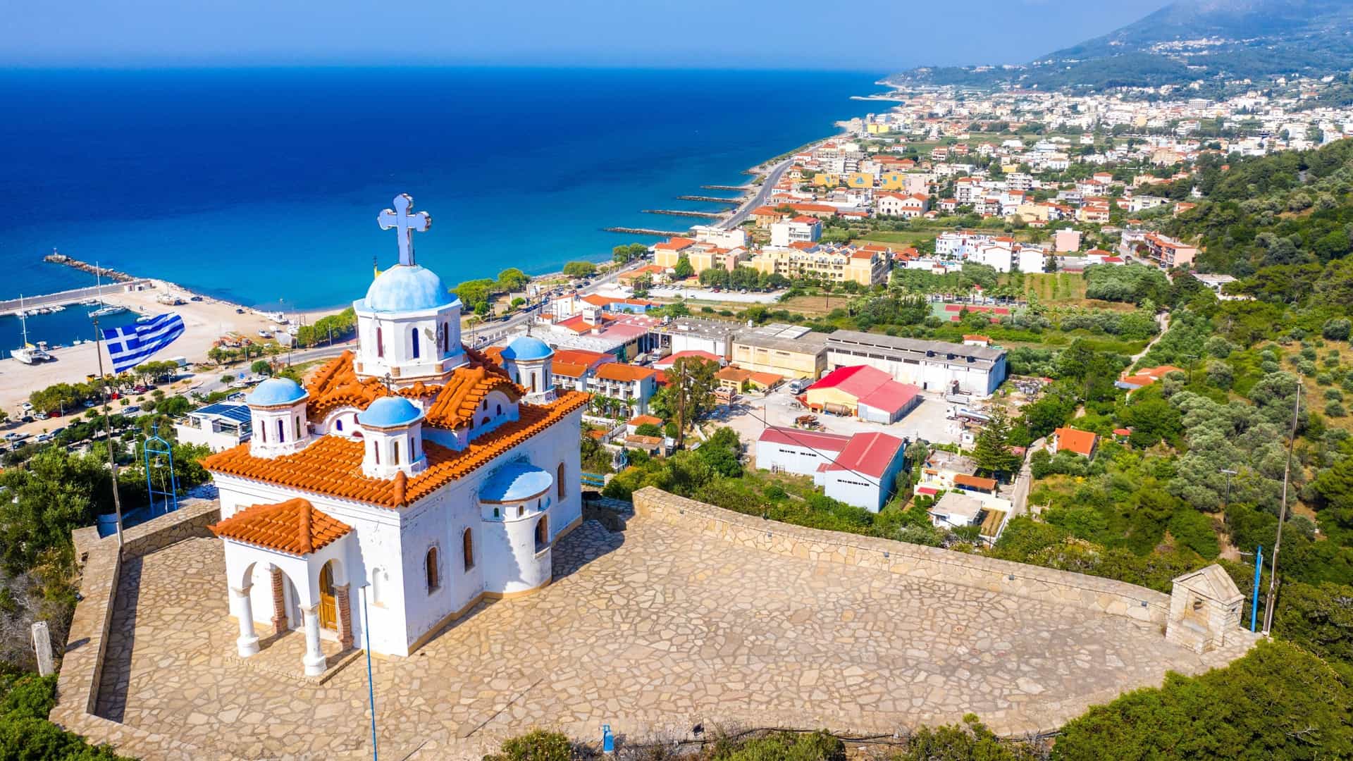 An aerial view of a white chapel with blue domes perched on a hillside, overlooking the coastal town of Samos and the deep blue Aegean Sea in Greece.