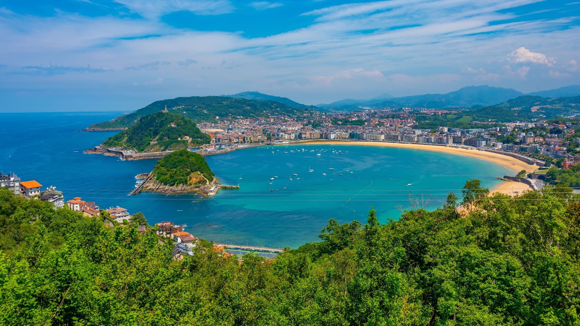A breathtaking aerial view of the Bay of La Concha and the city of San Sebastián, Spain, with the iconic crescent-shaped beach, boats in the turquoise water, and mountains in the background.