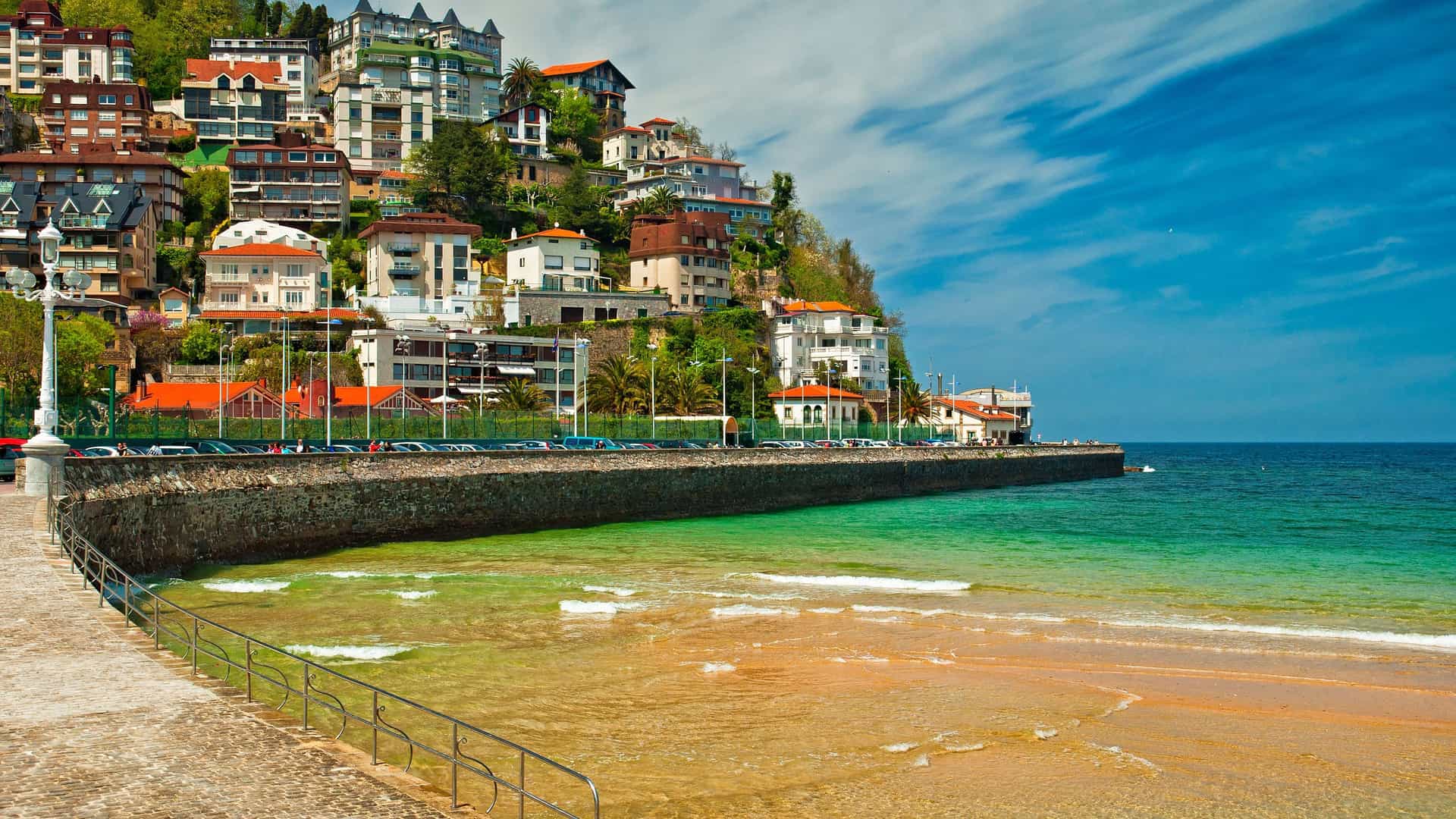 A view of a beautiful beach in San Sebastián, Spain, with a paved promenade and a stone sea wall, and a hillside covered with houses and hotels in the background.