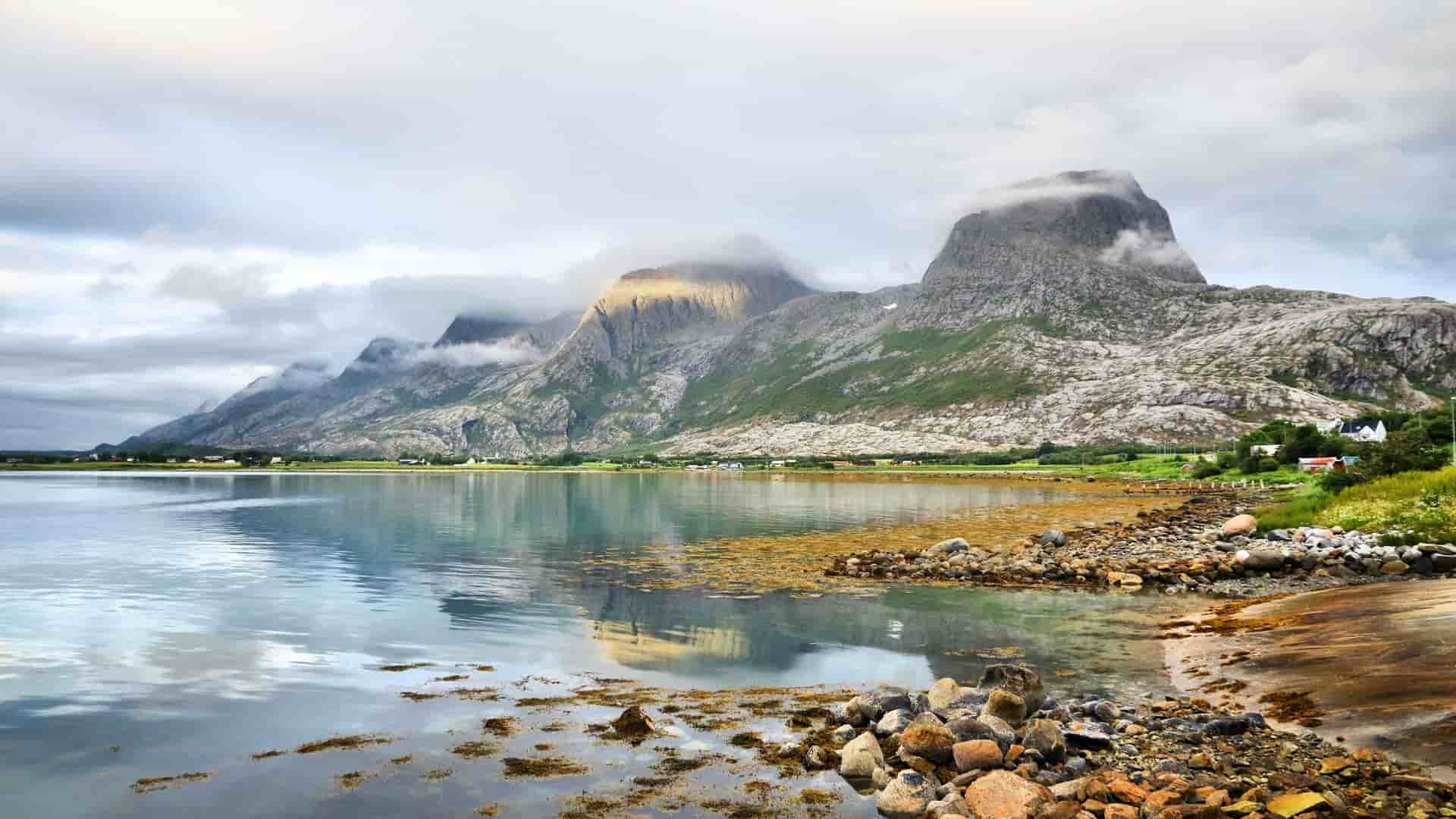 A serene landscape view of the Norwegian coastline in Sandnessjøen, with the Seven Sisters mountain range covered in low clouds reflecting on the still waters of the fjord.