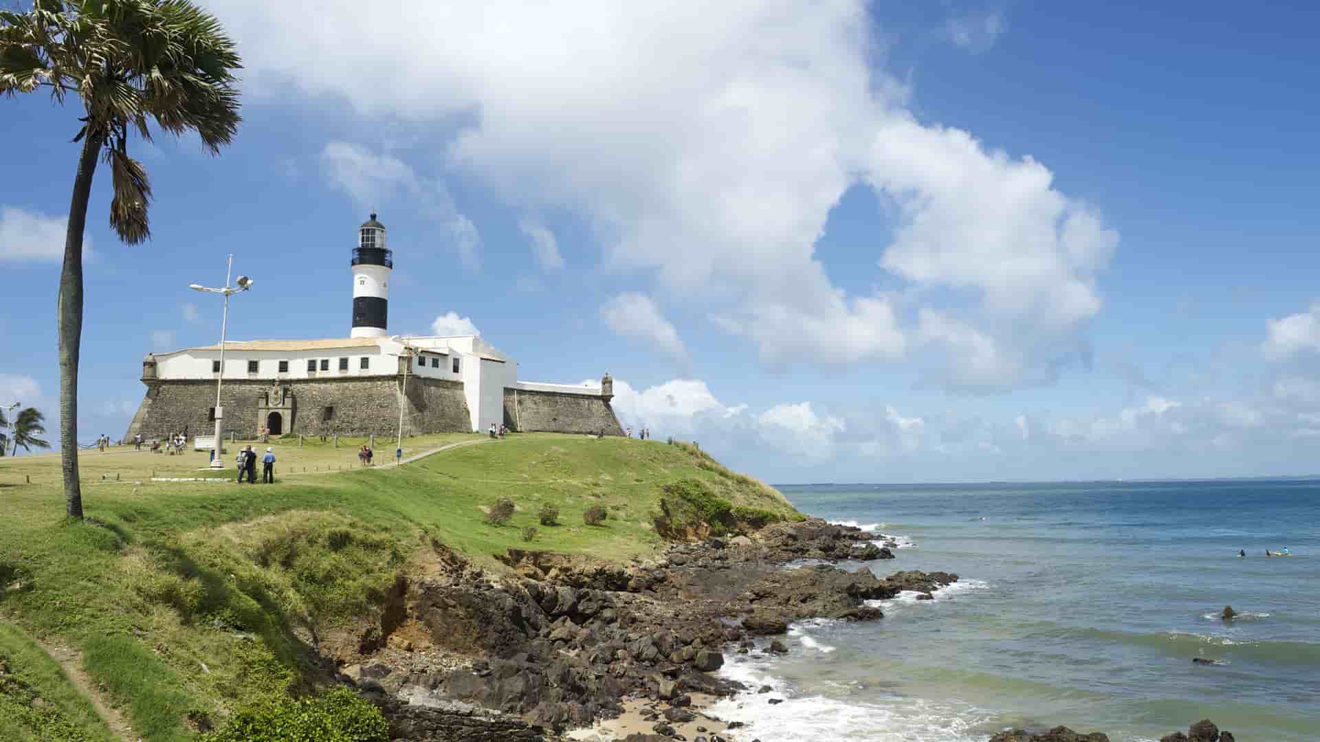 A beautiful seaside view of the Farol da Barra lighthouse and the historic Fort of Santo Antônio da Barra, a major landmark in Santarém, Brazil, with the Atlantic Ocean and a rocky coastline in the foreground.