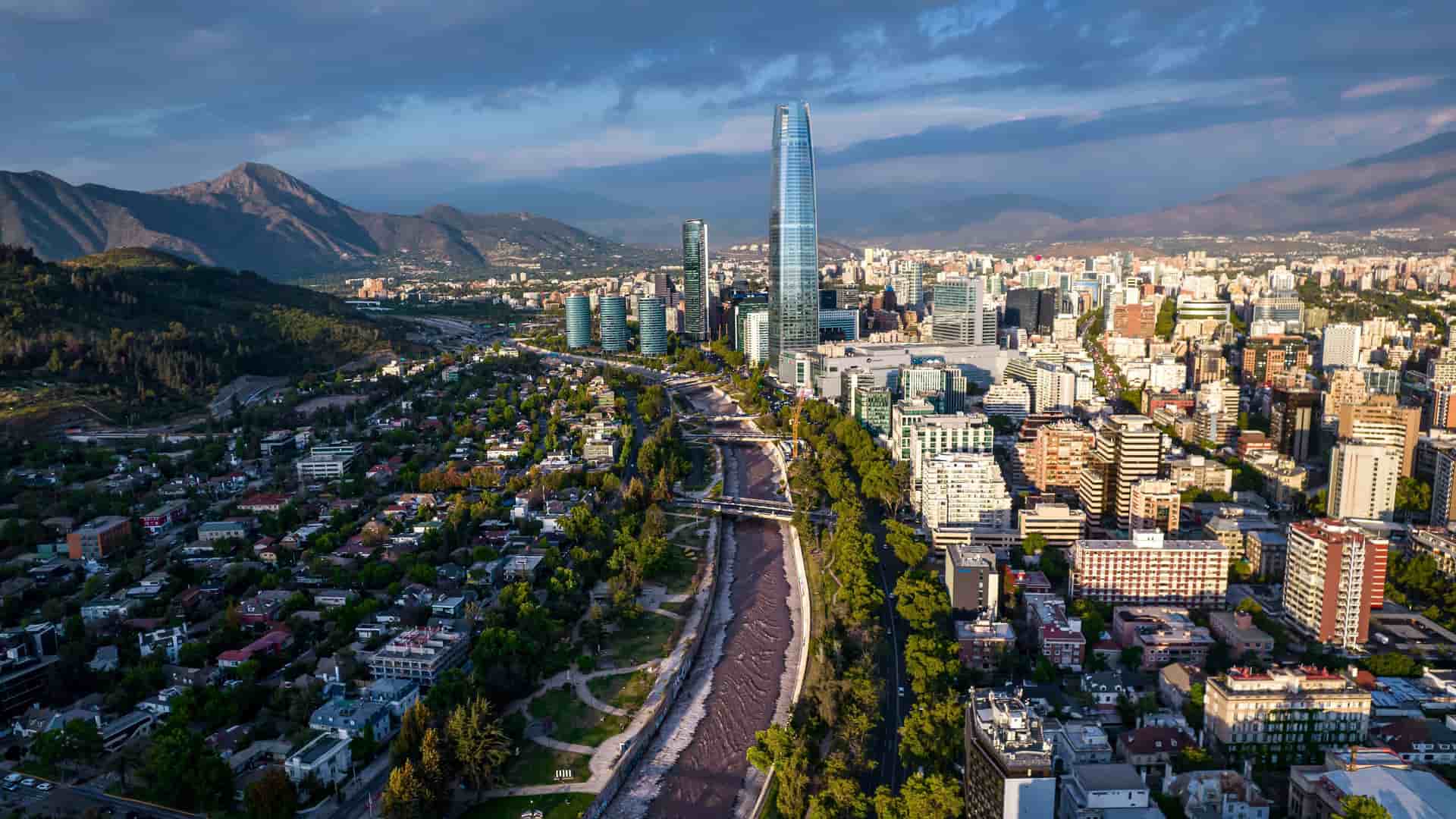An aerial panoramic view of the modern Santiago cityscape, with the Mapocho River flowing through it and the impressive Gran Torre Santiago skyscraper towering over the buildings, all set against the backdrop of the Andes Mountains.