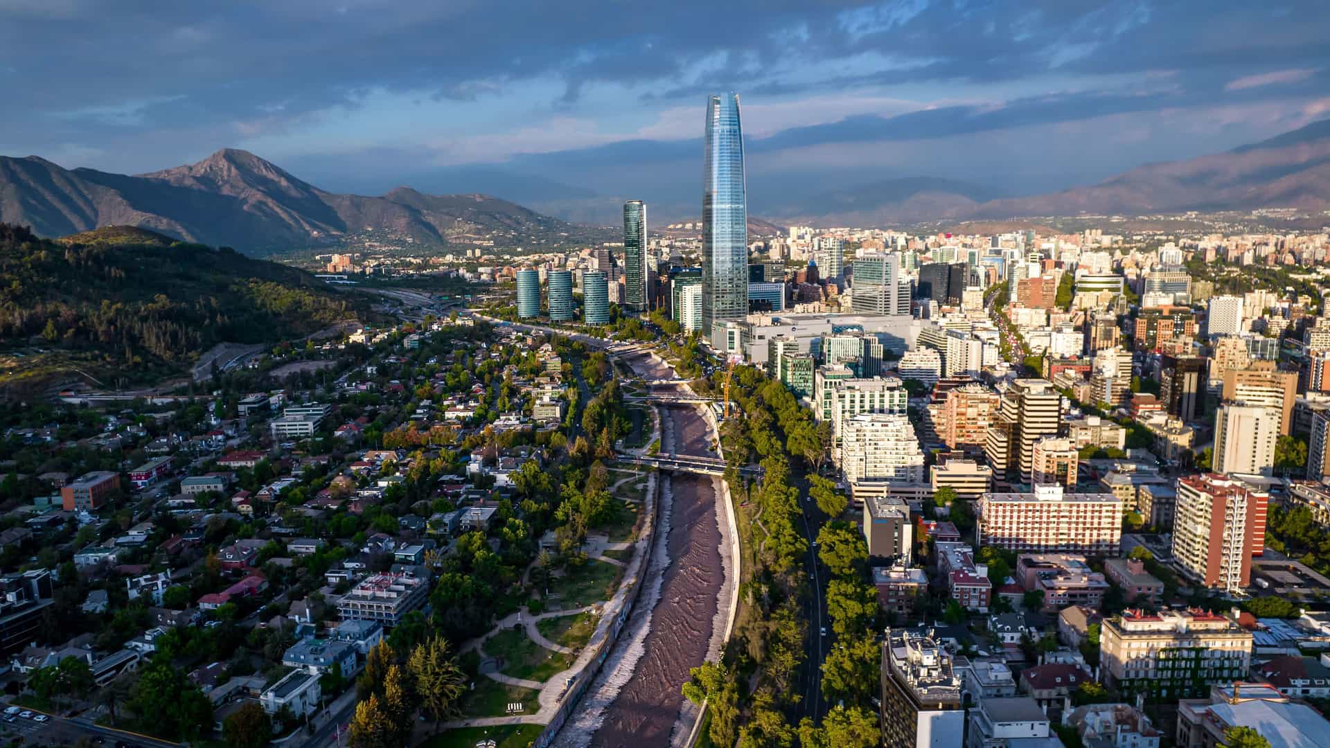 An aerial panoramic view of the modern Santiago cityscape, with the Mapocho River flowing through it and the impressive Gran Torre Santiago skyscraper towering over the buildings, all set against the backdrop of the Andes Mountains.