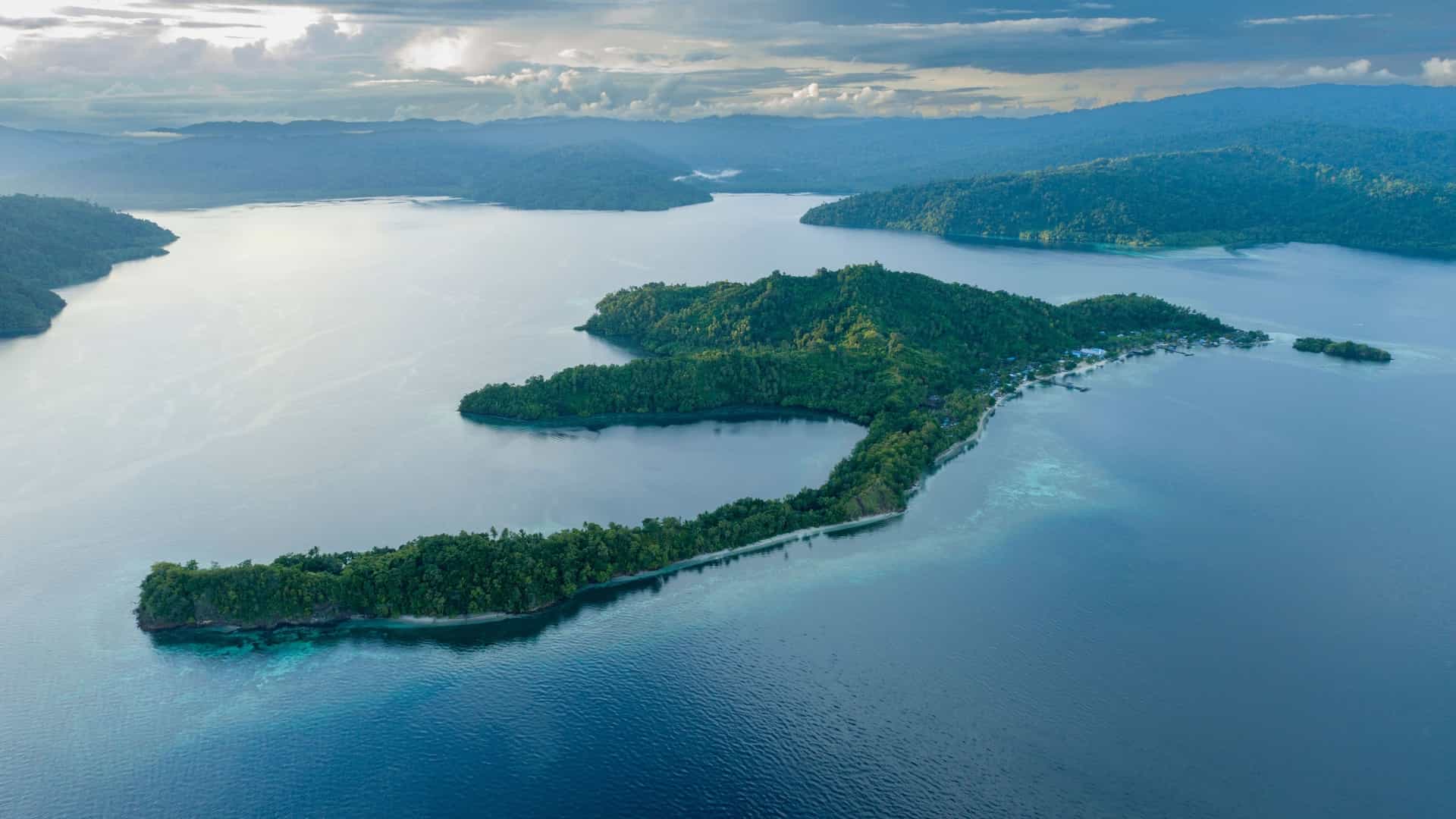 An aerial view of Sanwinggrai, a small island village in the Raja Ampat archipelago, with houses built on stilts over turquoise water and surrounded by lush green forests.